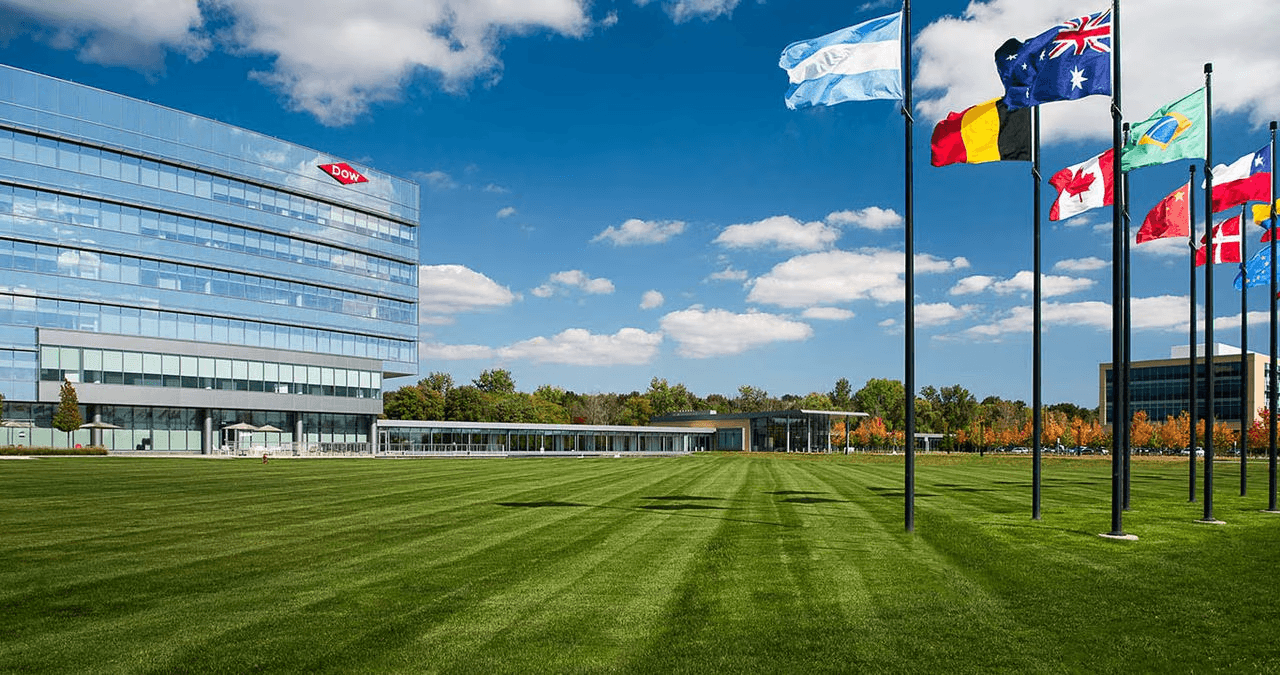 Modern office building with the Dow logo, surrounded by flagpoles with several national flags, on a bright, sunny day with a blue sky and scattered clouds.