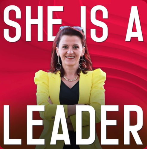 Woman smiling, wearing a yellow blazer and earrings, standing in front of a red background with white bold text that says 'SHE IS A LEADER'.