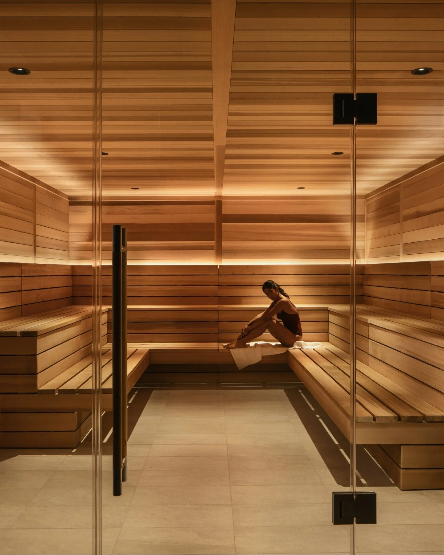 A woman sitting on a wooden bench inside a modern sauna room, illuminated with warm lighting.