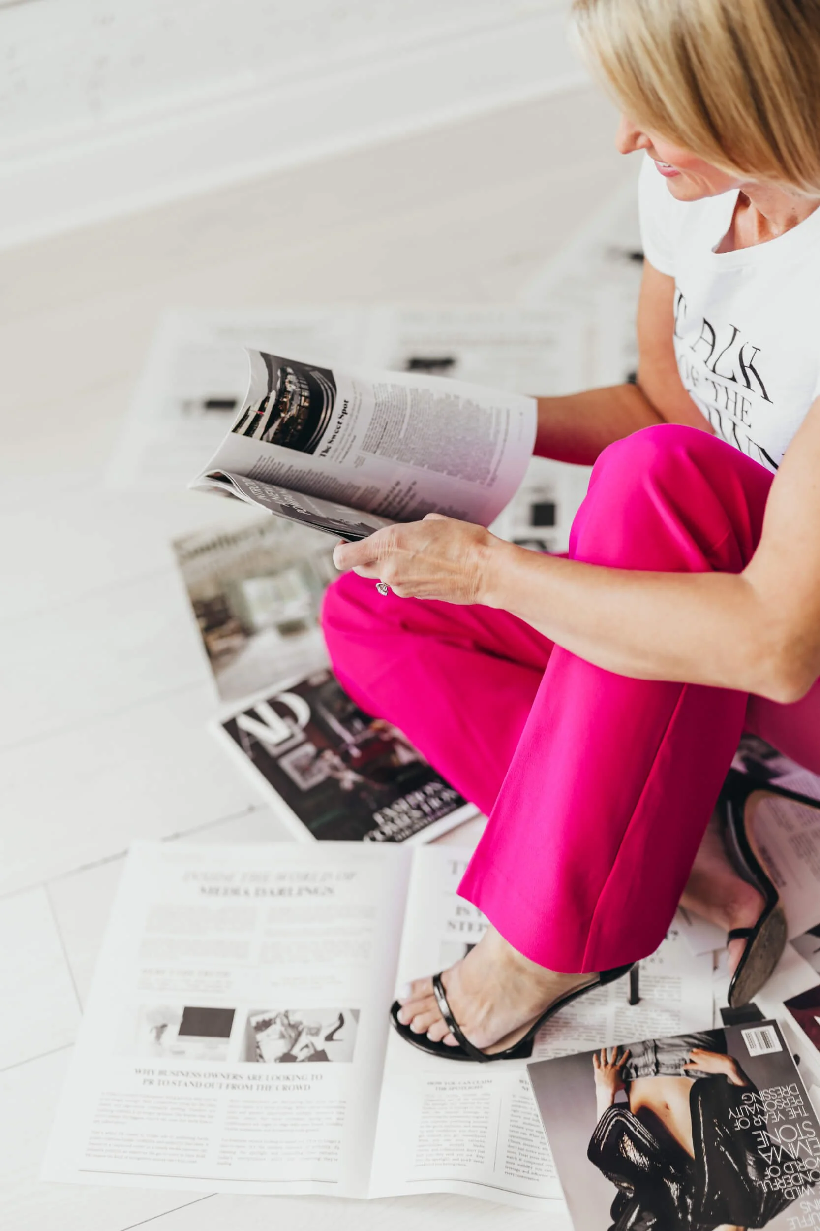 A woman in pink pants and black sandals is seated on a chair, reading a magazine. Various magazines and printed newspapers are spread on the white floor around her.