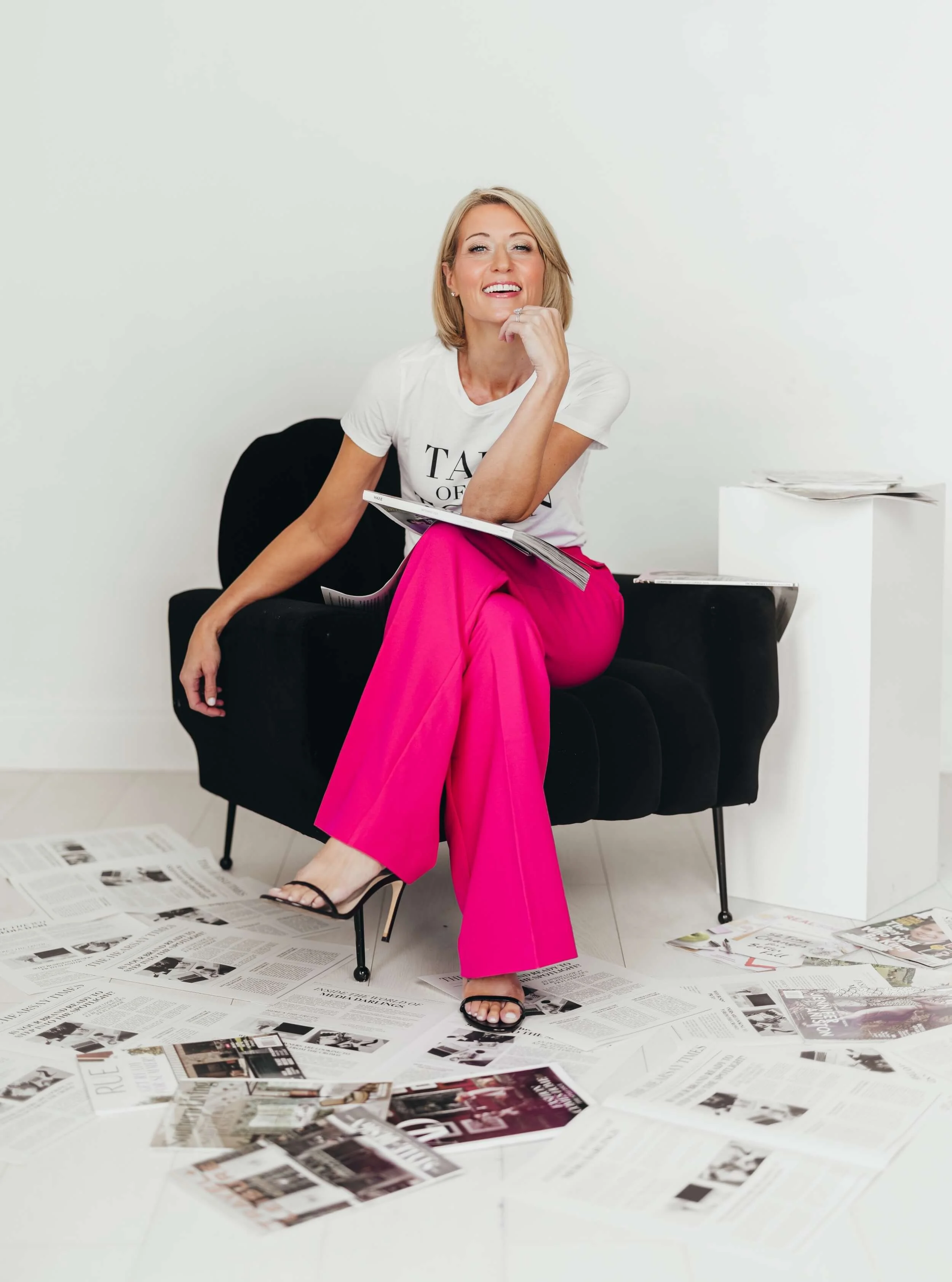 Woman sitting on a black sofa with scattered newspapers on the floor and on a side table, smiling at the camera, wearing a white t-shirt, bright pink pants, and black sandals, in a minimal white room.