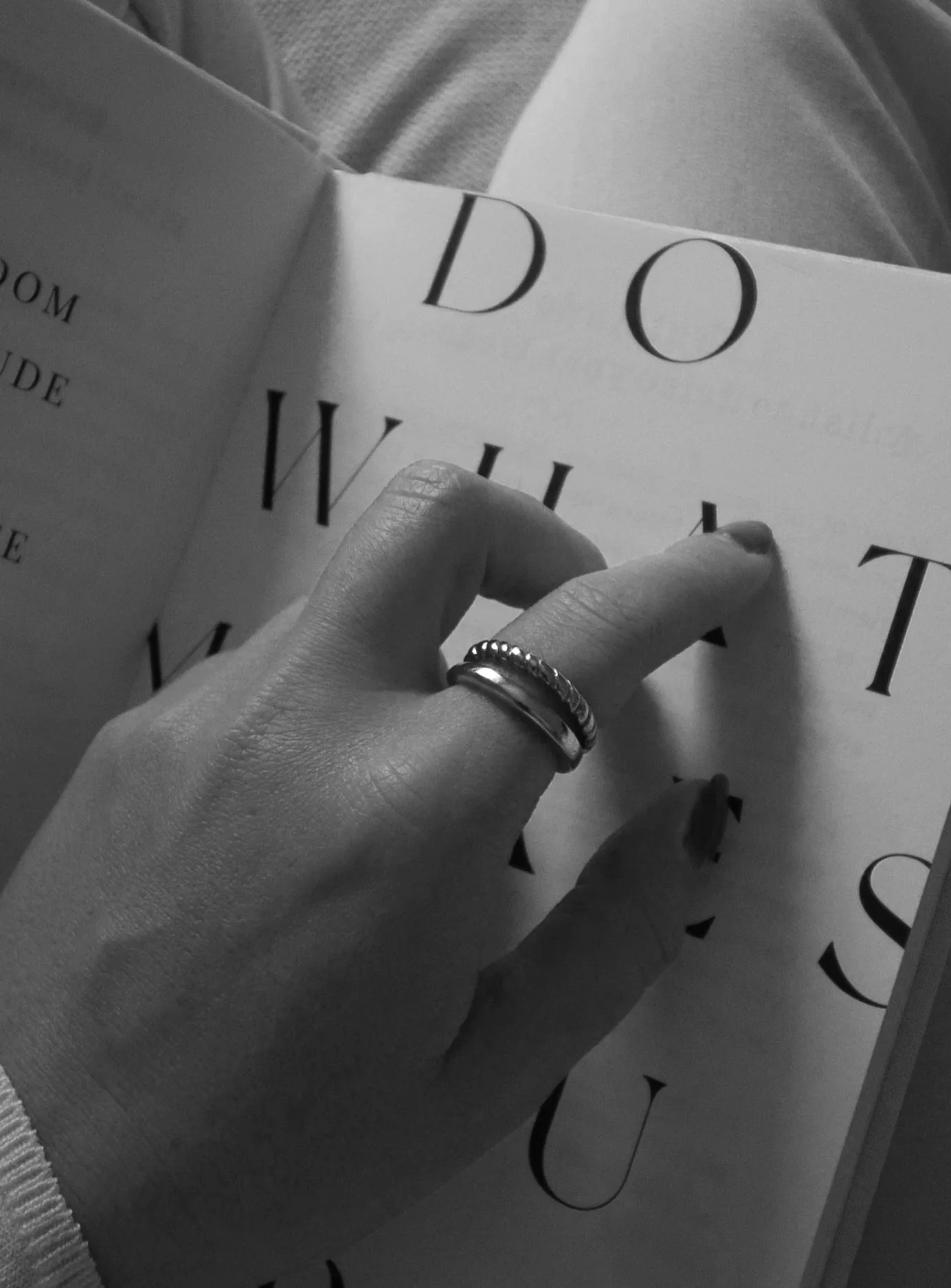 A person with rings on their fingers is flipping through pages of a book or magazine with large, bold lettered text, sitting on a surface with decorative black swirl patterns.