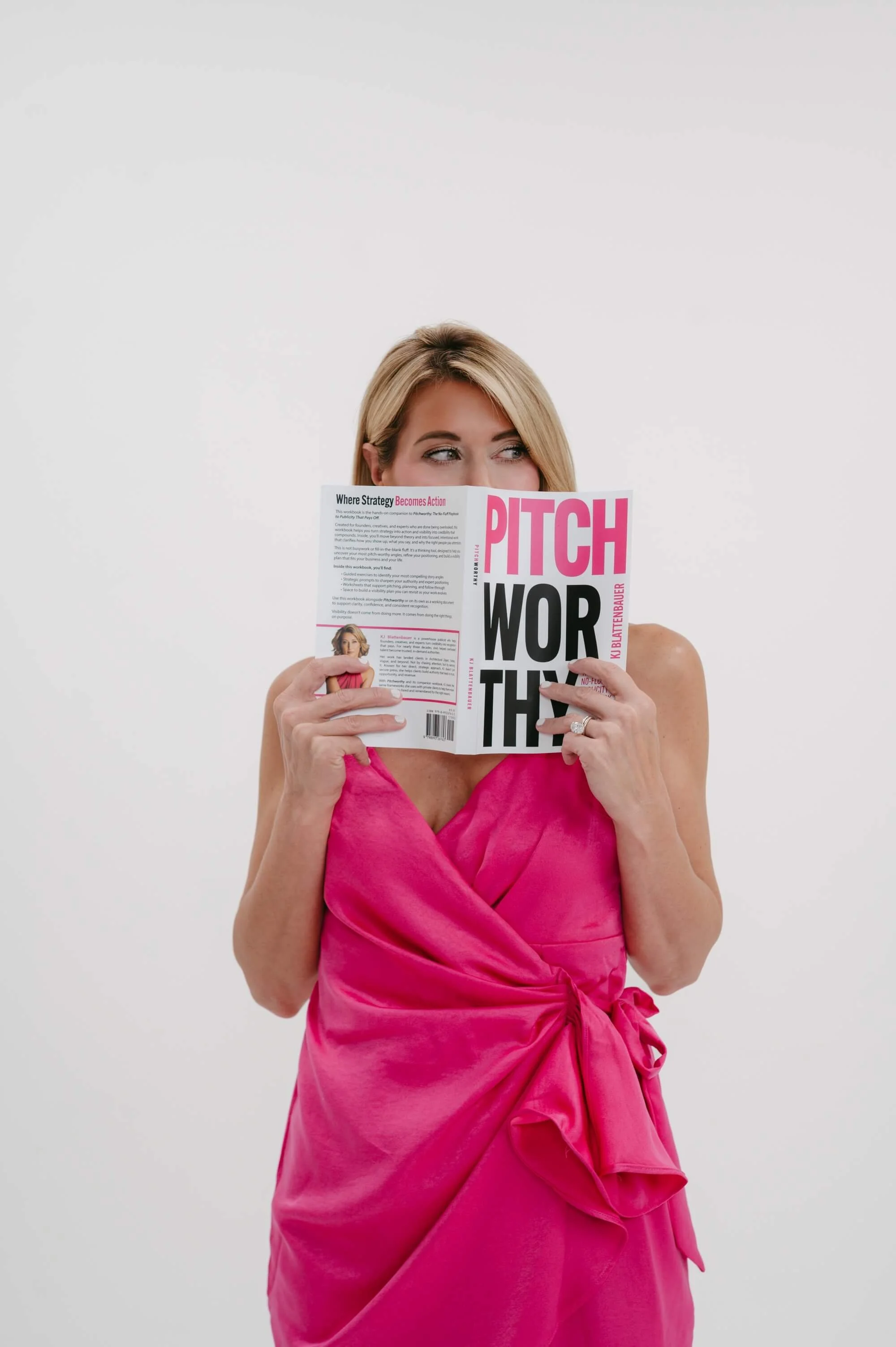 A woman wearing a pink dress, holding a magazine titled 'Pitch' in front of her face, with only her eyes and part of her head visible, against a plain white background.