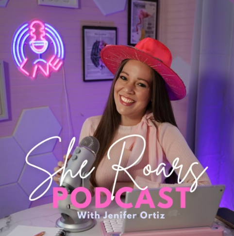 A woman with long dark hair wearing a red wide-brimmed hat smiling at the camera, sitting at a desk with a microphone, laptop, and pink notebook in front of her, in a room decorated with neon signs and framed pictures.