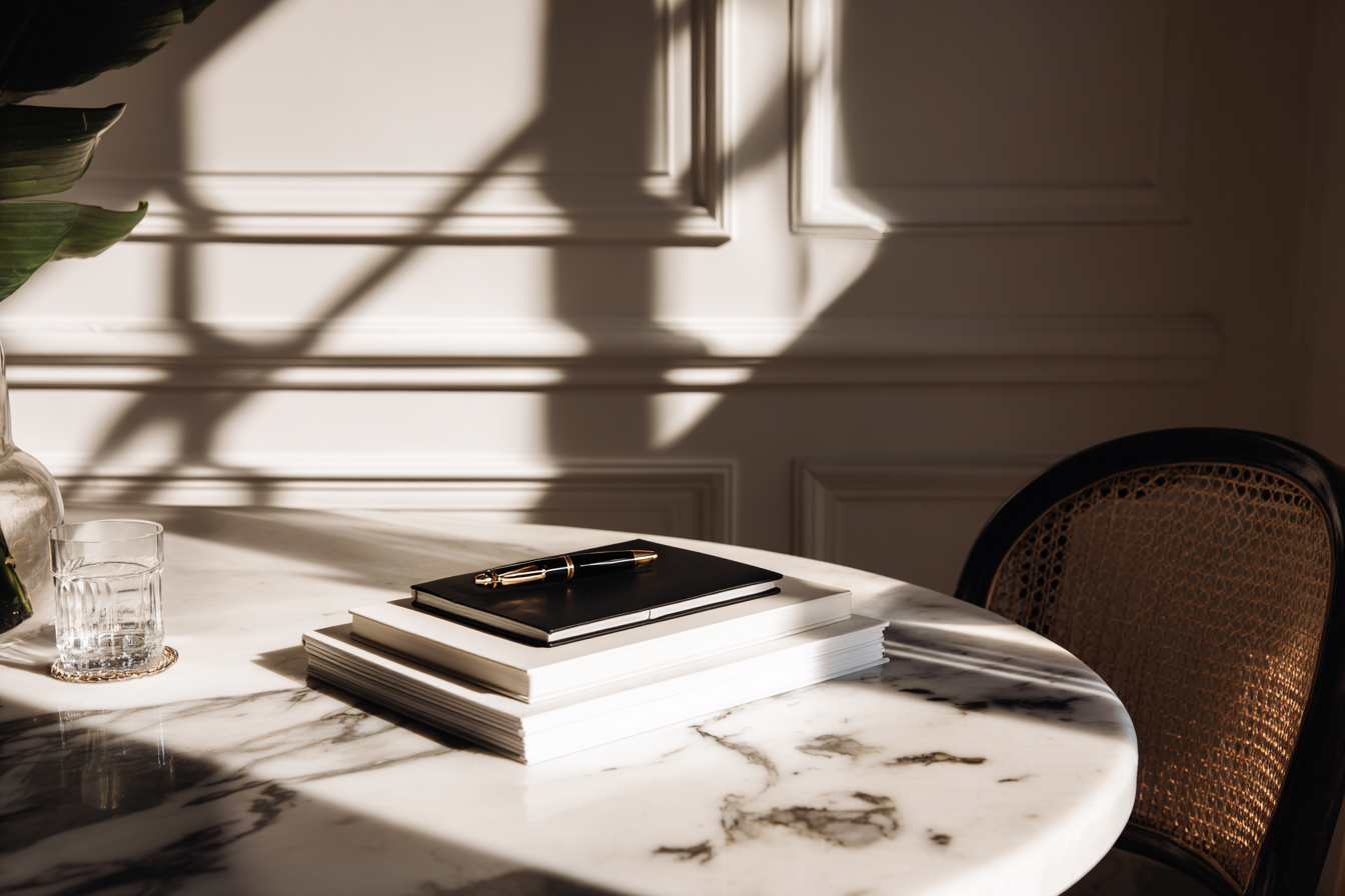 Sunlight casts shadows on a white marble table with a stack of notebooks, a pen, a glass of water, and a plant in a clear vase, next to a black chair with a woven cane backrest.