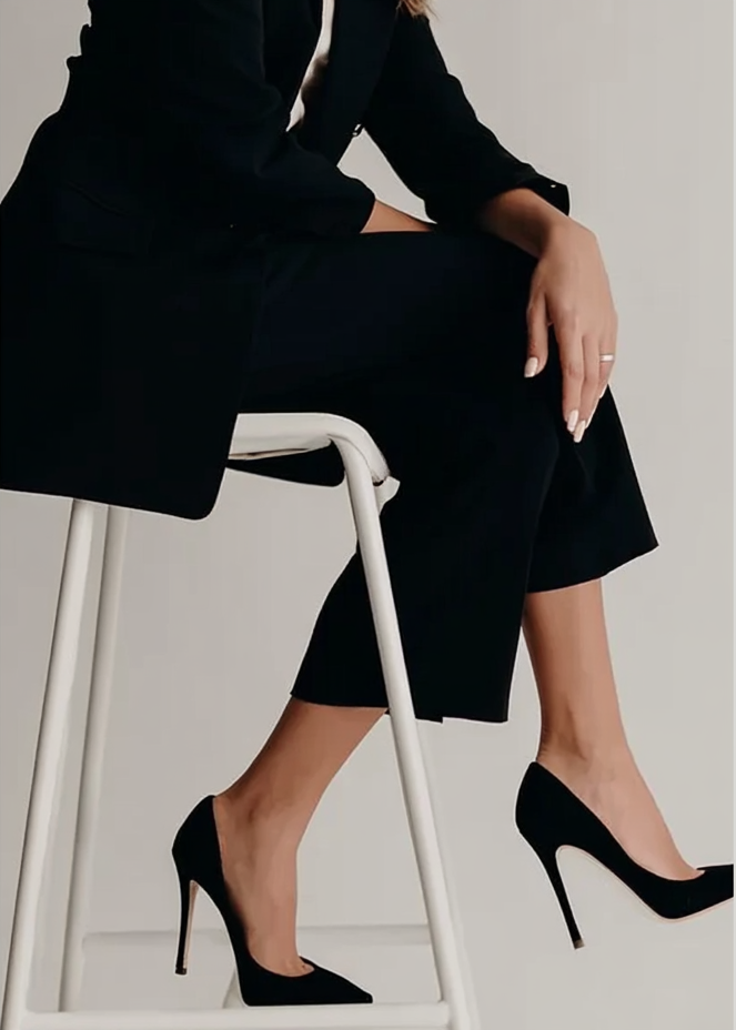 A woman in black clothing sitting on a white chair, with her legs crossed and wearing black high heels.