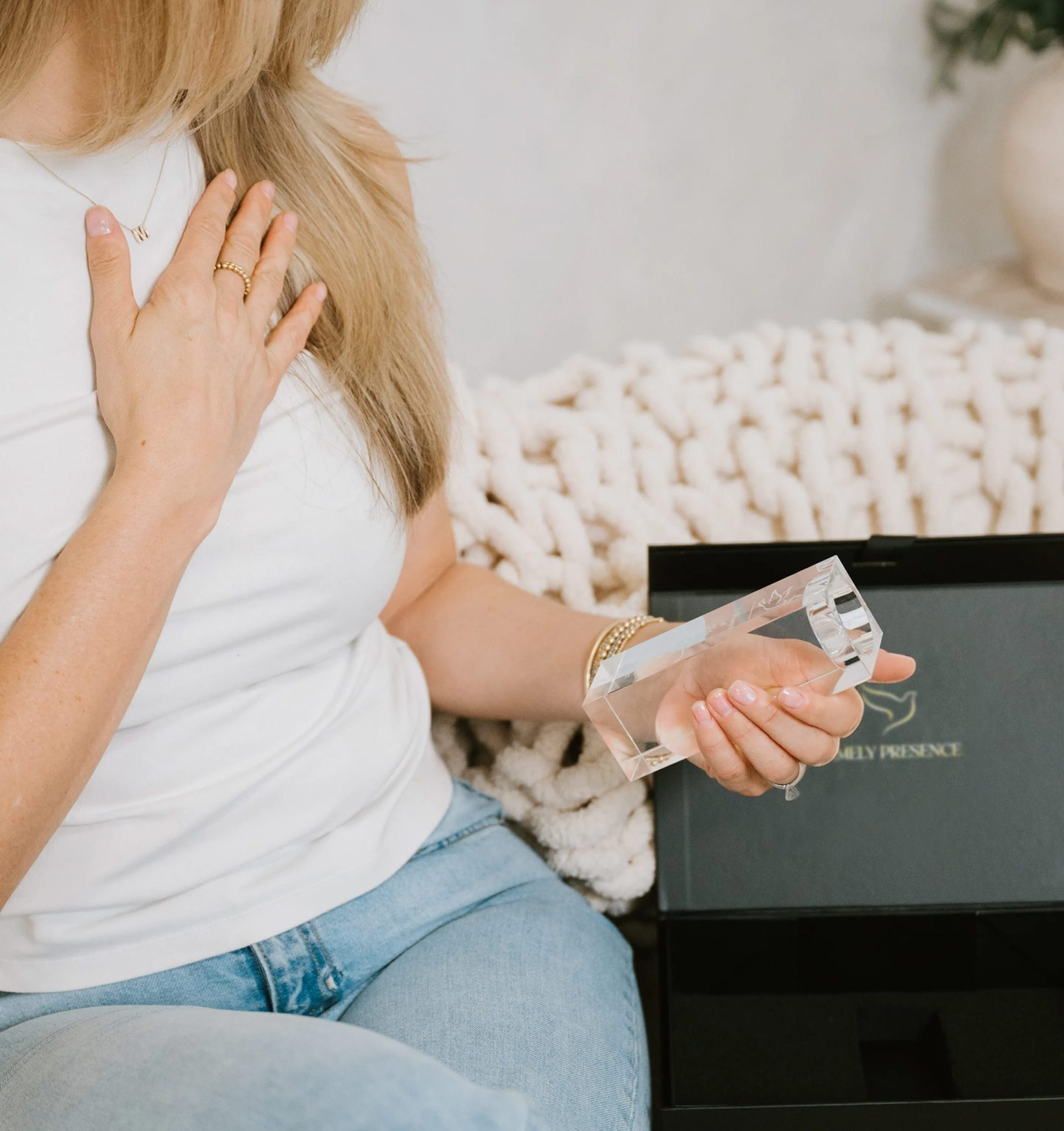 Woman wearing a white top and blue jeans sitting on a beige plush rug, holding a clear glass award or trophy, with a black display box and plant in the background.