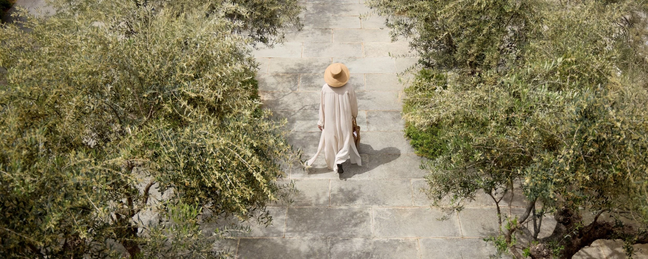 A woman dressed in a flowing white dress and a large straw hat walks on a stone pathway surrounded by green bushes.