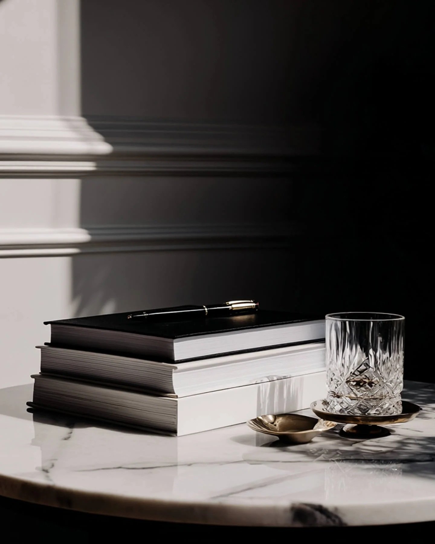 A stack of black and white books on a marble table with a glass of whiskey, a silver spoon, and a silver tray, with sunlight casting shadows.