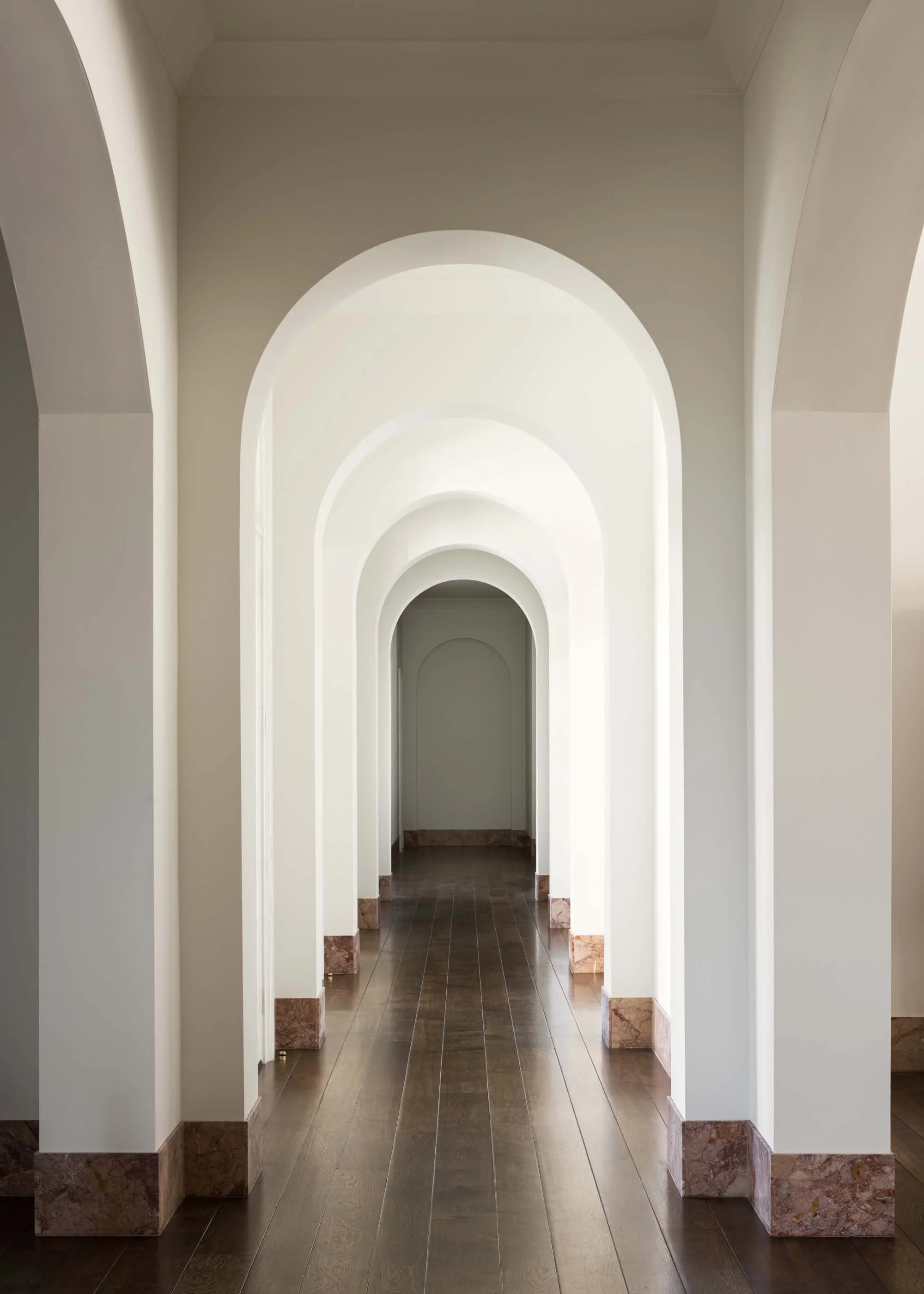 A long corridor with multiple white archways and brown tiled flooring, leading to a door at the end.