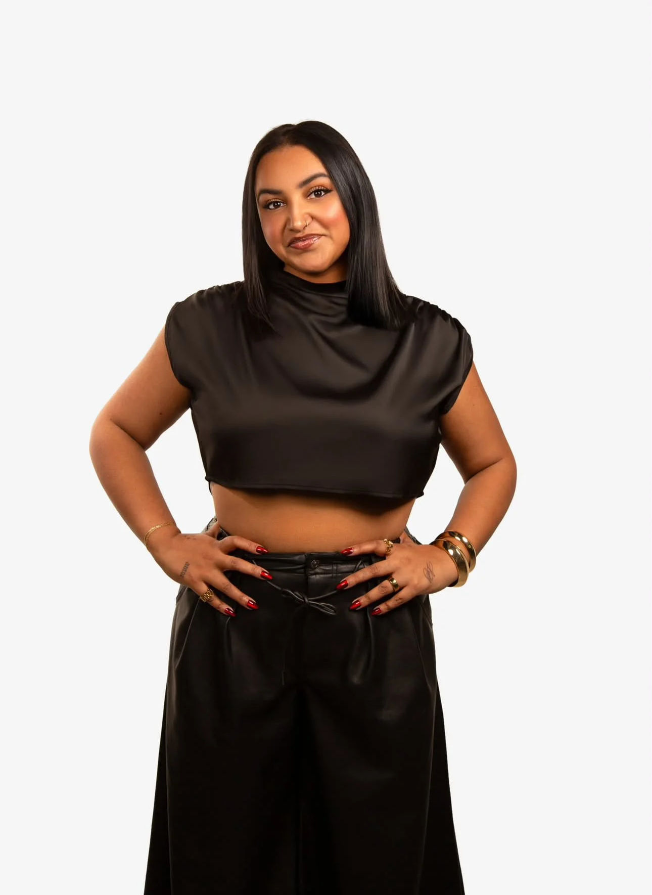 A woman with medium skin tone and black hair posing with her hands on her hips, wearing a black satin crop top and black pants, standing against a plain white background.