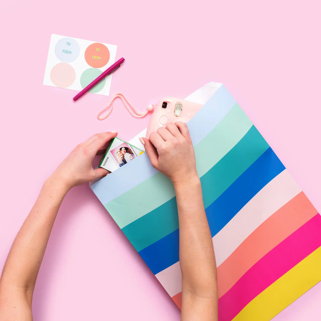 Person wrapping a gift box with rainbow stripes. Supplies include a pink camera, a colorful 'To' and 'From' sticker sheet, and a pink marker on a pink background.
