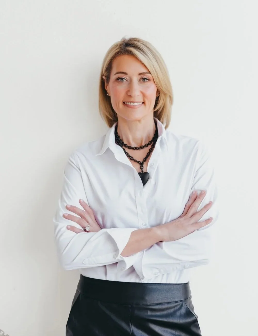A woman with blonde hair wearing a white shirt, black leather pants, and layered black necklaces, standing with arms crossed against a plain light-colored wall.
