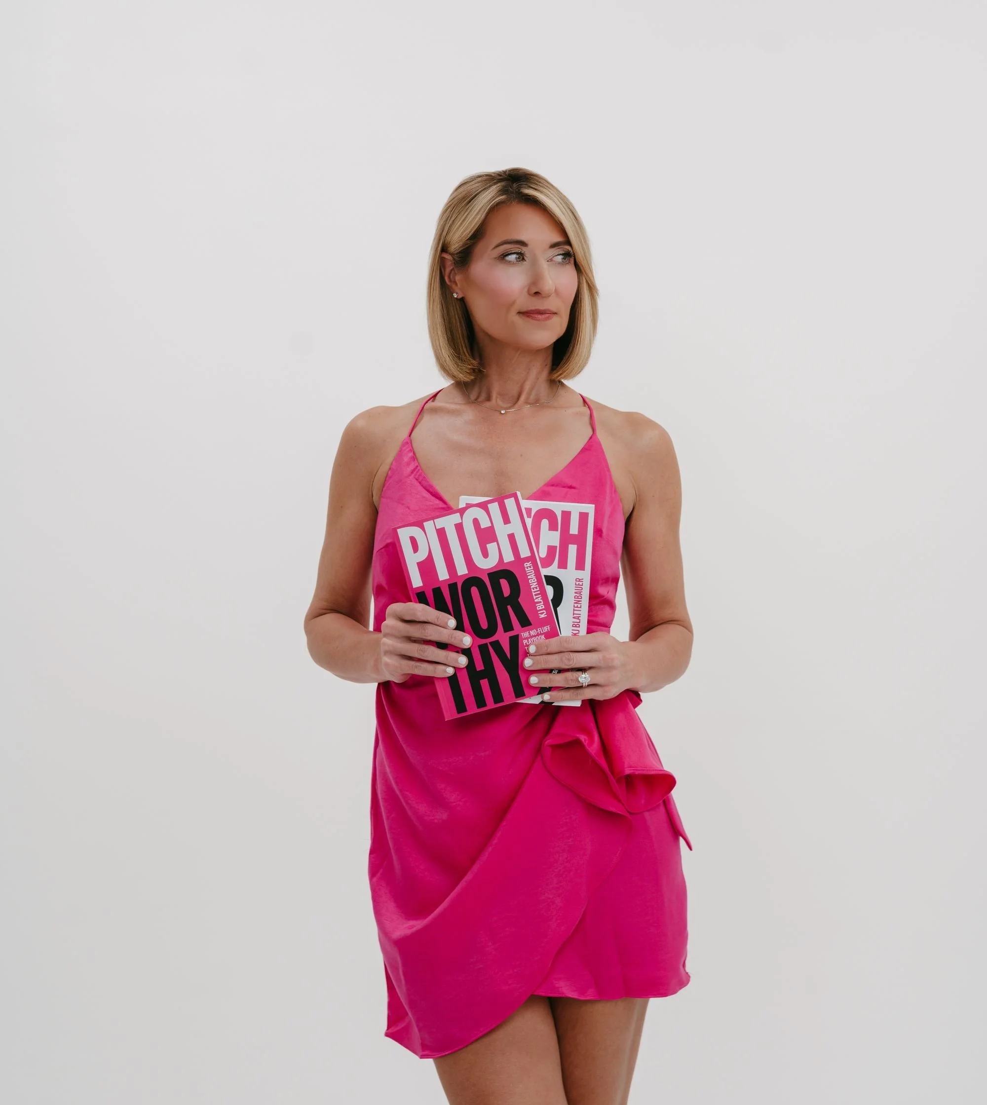 Woman with shoulder-length blonde hair wearing a pink dress and holding a pink card, standing against a plain white background.