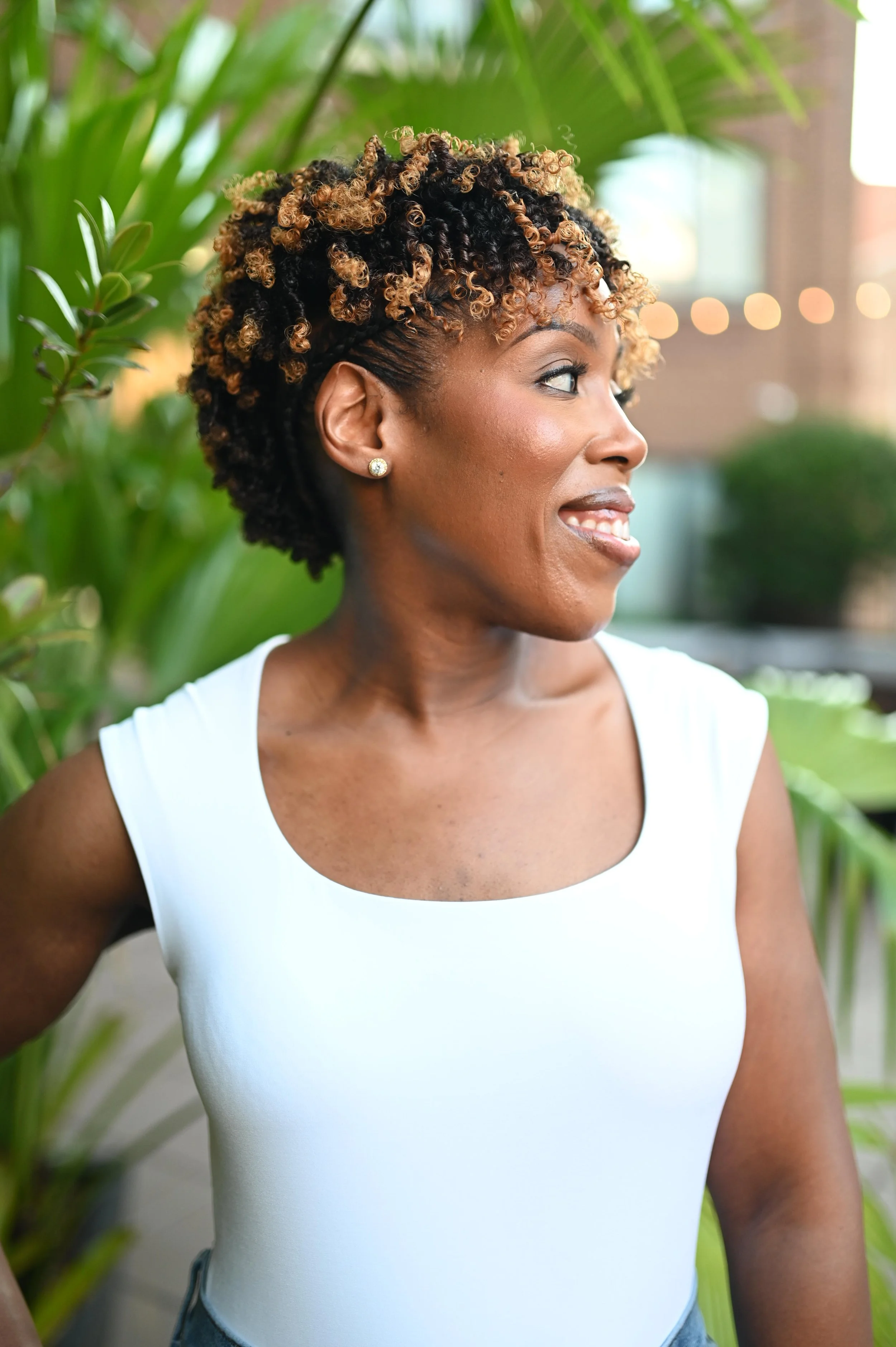 Profile of a woman with curly, short hairstyle with black and blonde curls, wearing pearl earrings and a white sleeveless top, standing outdoors with greenery in the background.