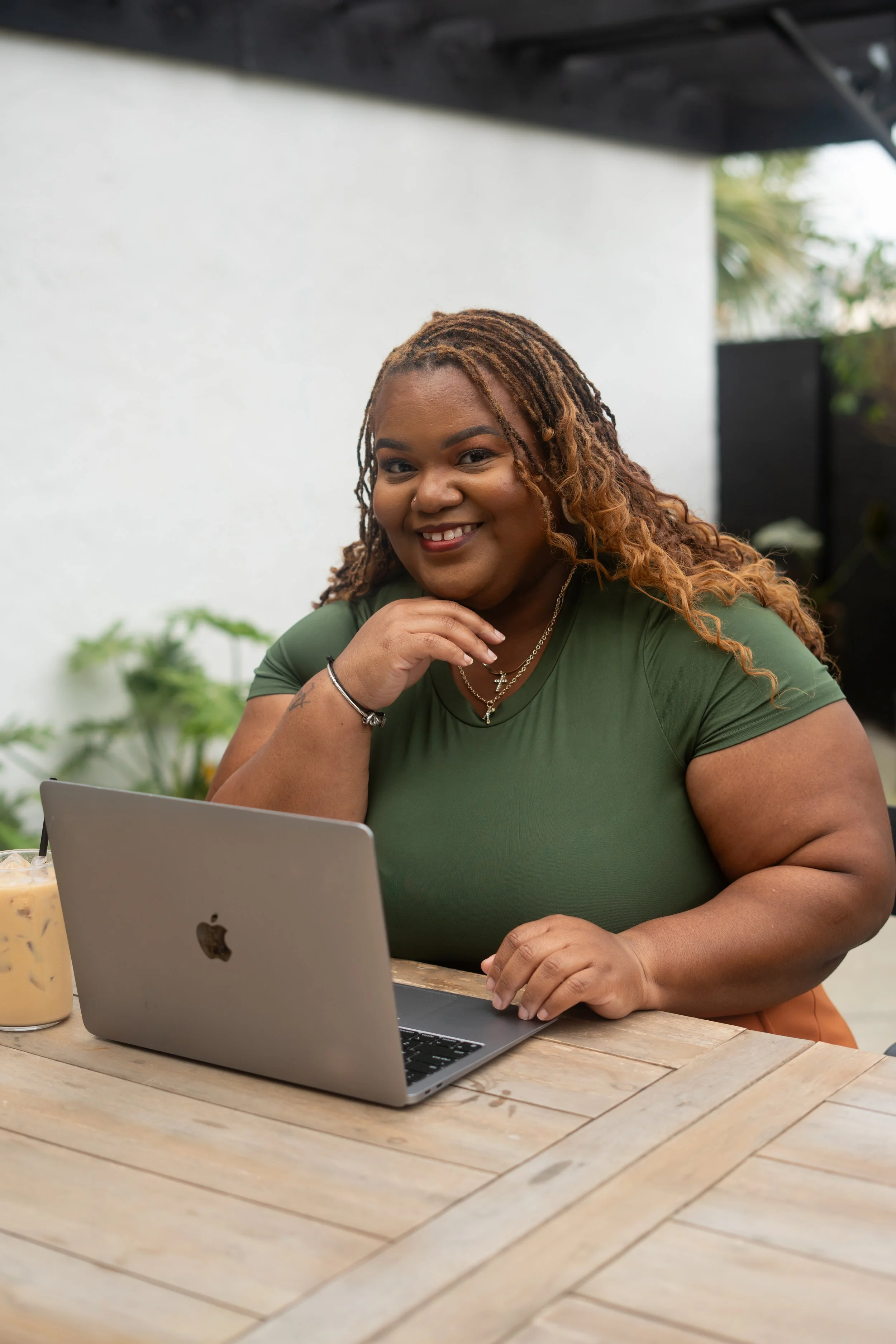 A woman with curly hair, wearing an olive green shirt, sitting at a wooden table with a silver laptop and a glass of iced coffee, smiling at the camera.