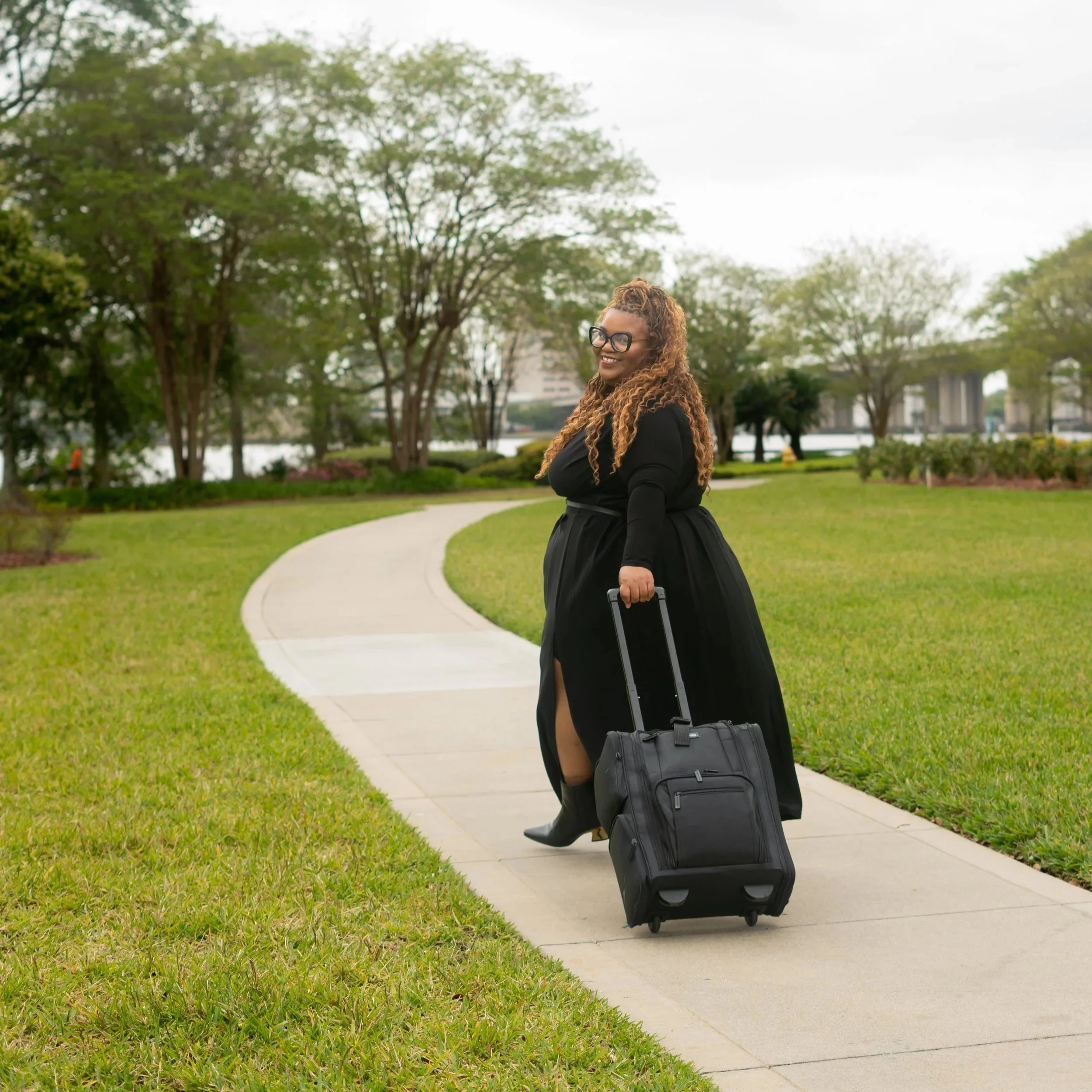 Woman with curly hair and glasses smiling, walking in a park with a rolling suitcase, trees, and a path visible in the background.