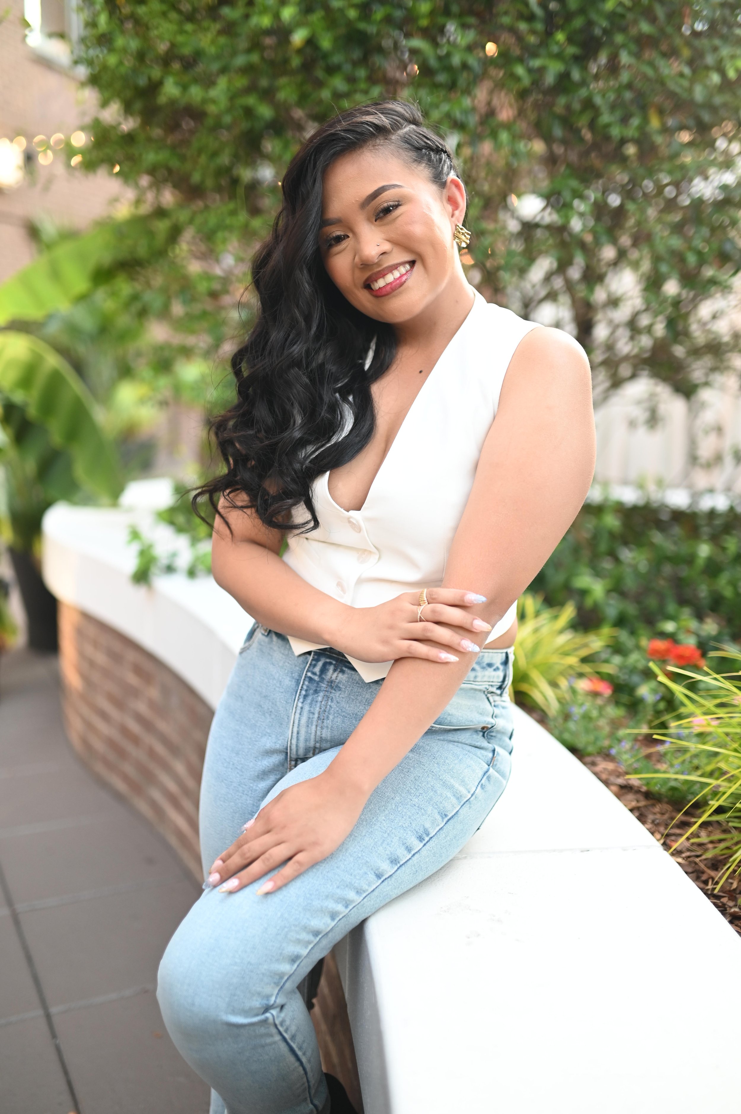A woman with long, curly black hair, wearing a white sleeveless top and light blue jeans, sitting on a white ledge outdoors, smiling with her arms crossed. Background features green foliage and a brick wall.