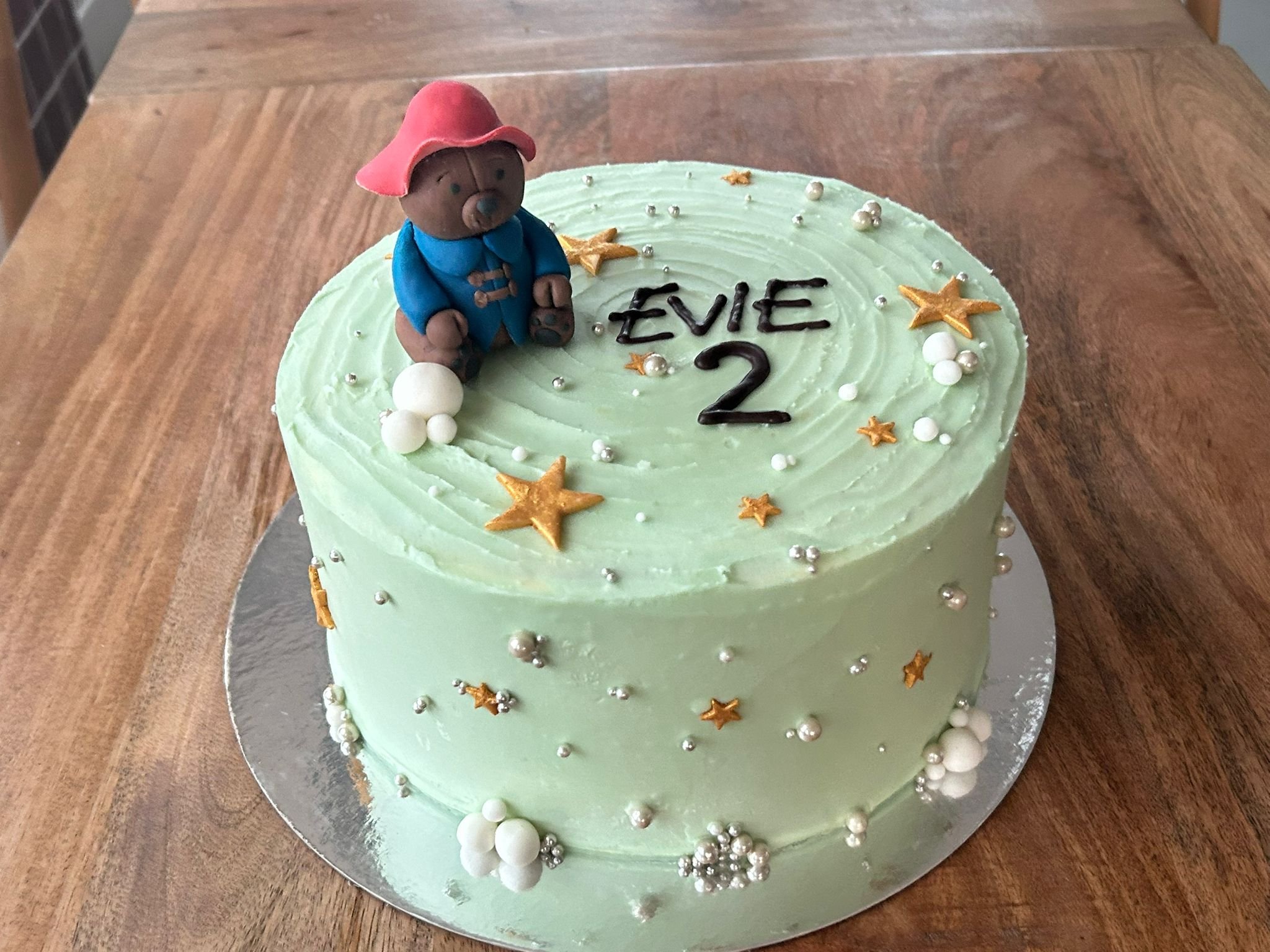 A round birthday cake with mint green frosting, decorated with gold stars, white and silver pearls, and a fondant bear wearing a pink hat and blue coat sitting on top. The cake displays the name 'Evie' and the number '2' for a second birthday celebration.