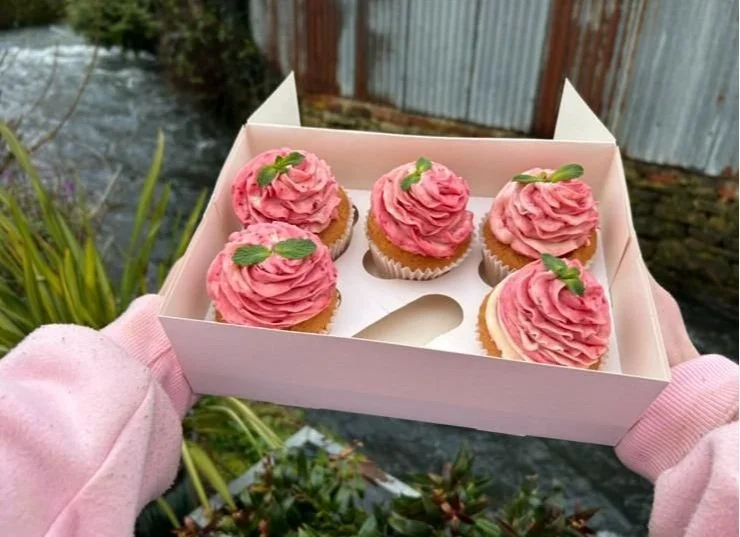 A person wearing pink gloves holding a white bakery box with six cupcakes decorated with pink swirled frosting and small green garnish, outdoors in a garden setting.
