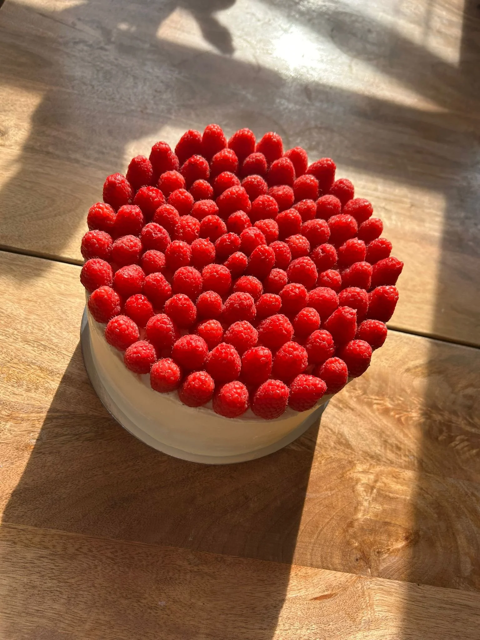 A round cake topped with fresh raspberries, placed on a wooden surface with sunlight casting shadows.