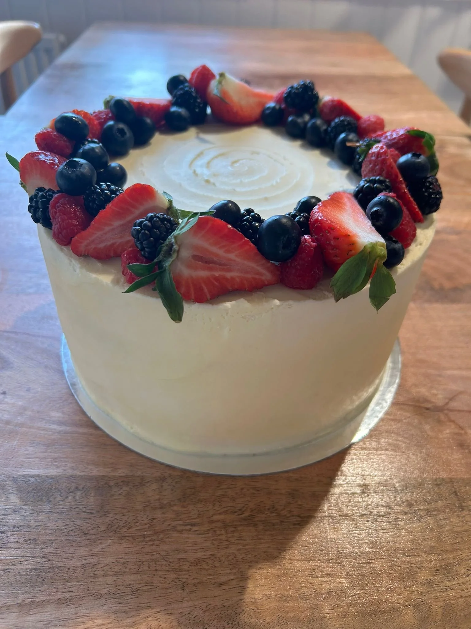 A round cake with white frosting decorated with sliced strawberries, blueberries, blackberries, and some green leaves on top, placed on a wooden table.