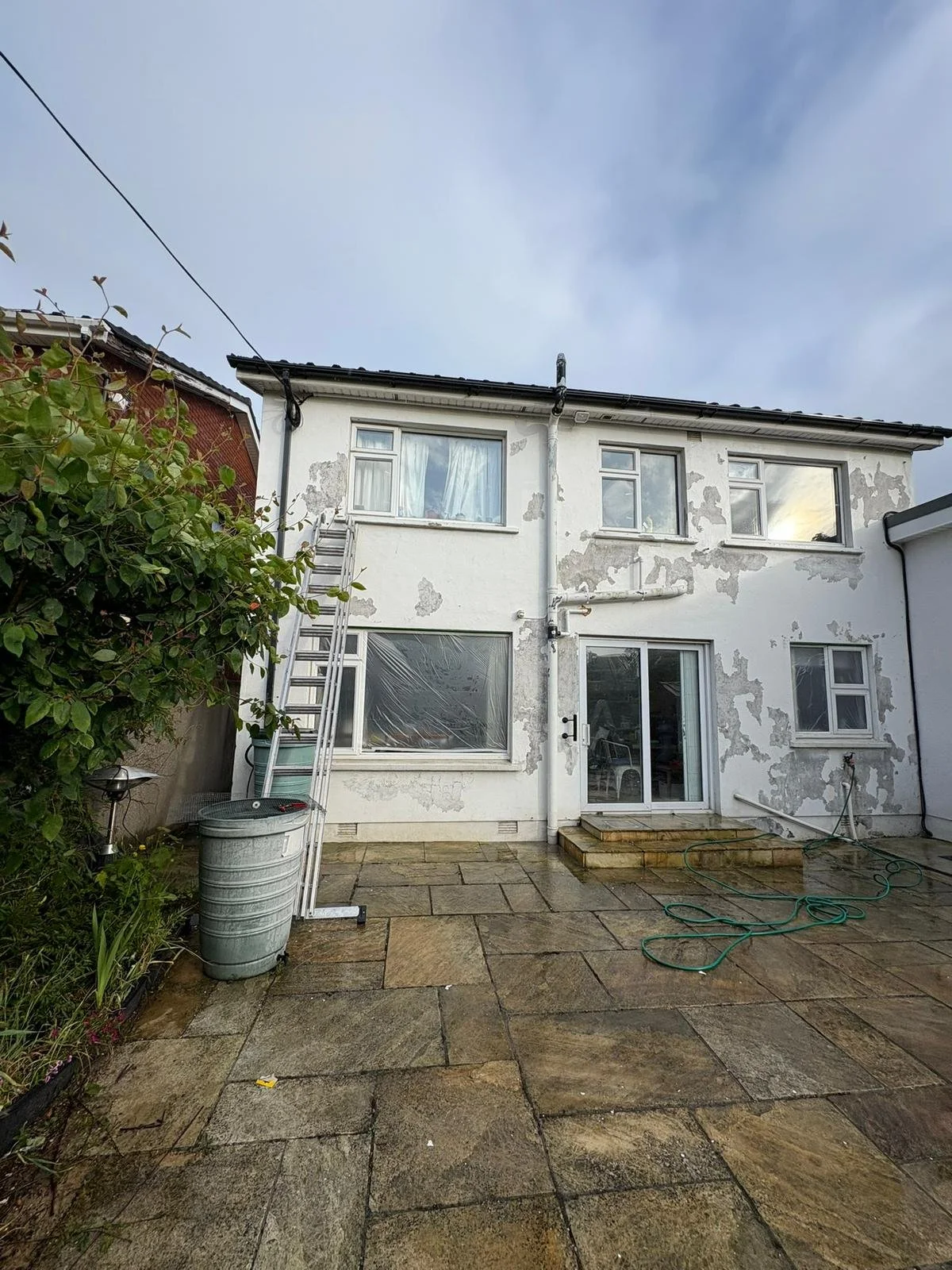 Back of a white house with peeling paint, a glass sliding door, and four windows on the upper level. There is an outdoor patio with wet paving stones, a garden hose, a ladder, a trash bin, and some greenery on the left side.