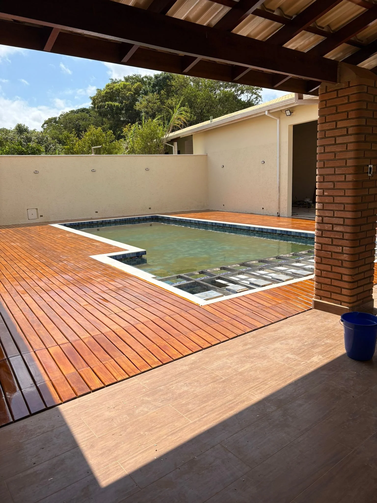 View of a backyard with a wooden deck, a small swimming pool under construction, a white wall, and lush green trees in the background.