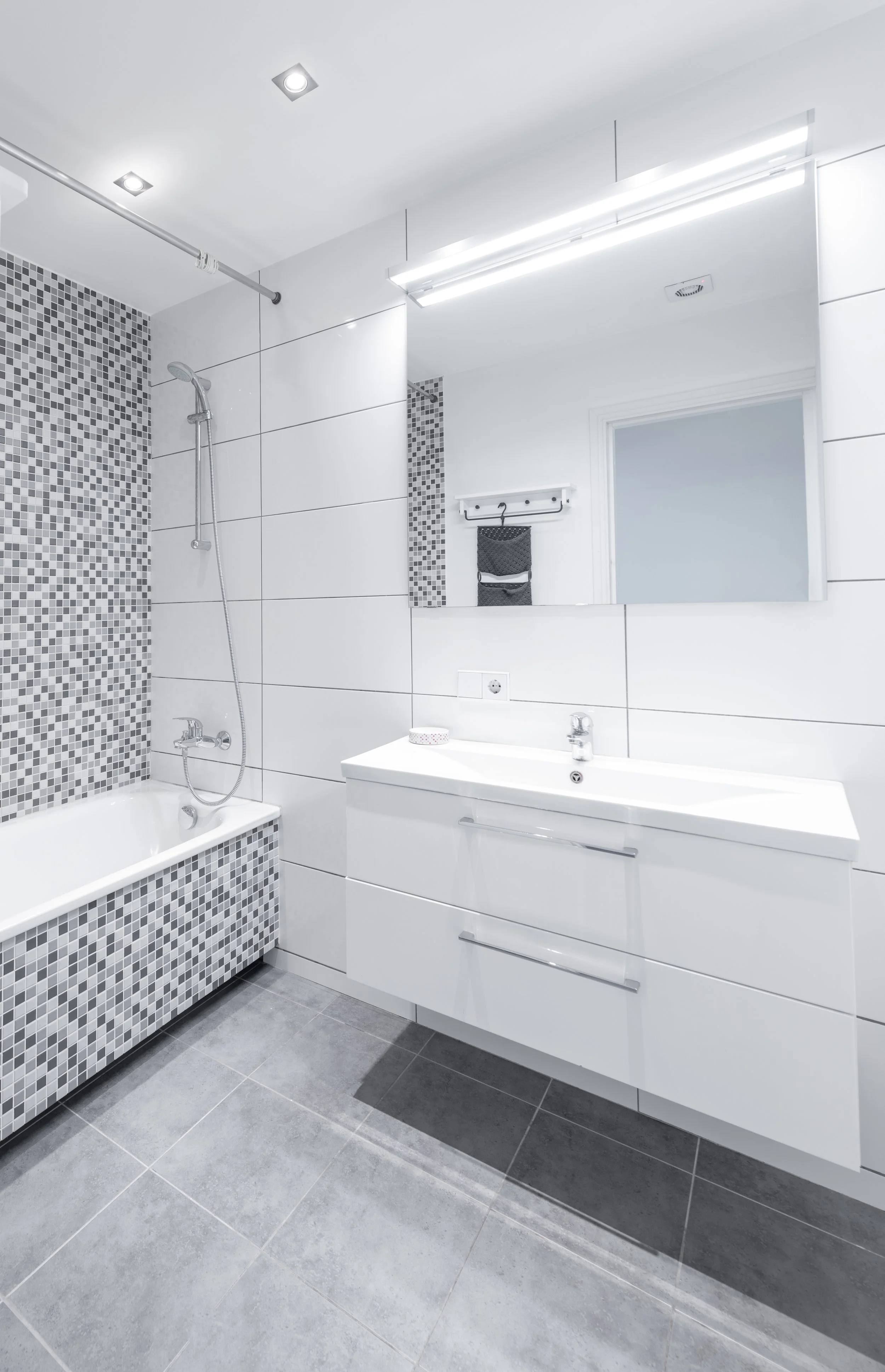 Modern white bathroom with a bathtub on the left, gray and white mosaic tile accent wall, a white floating vanity with a sink and drawers, a large mirror, and overhead lighting.