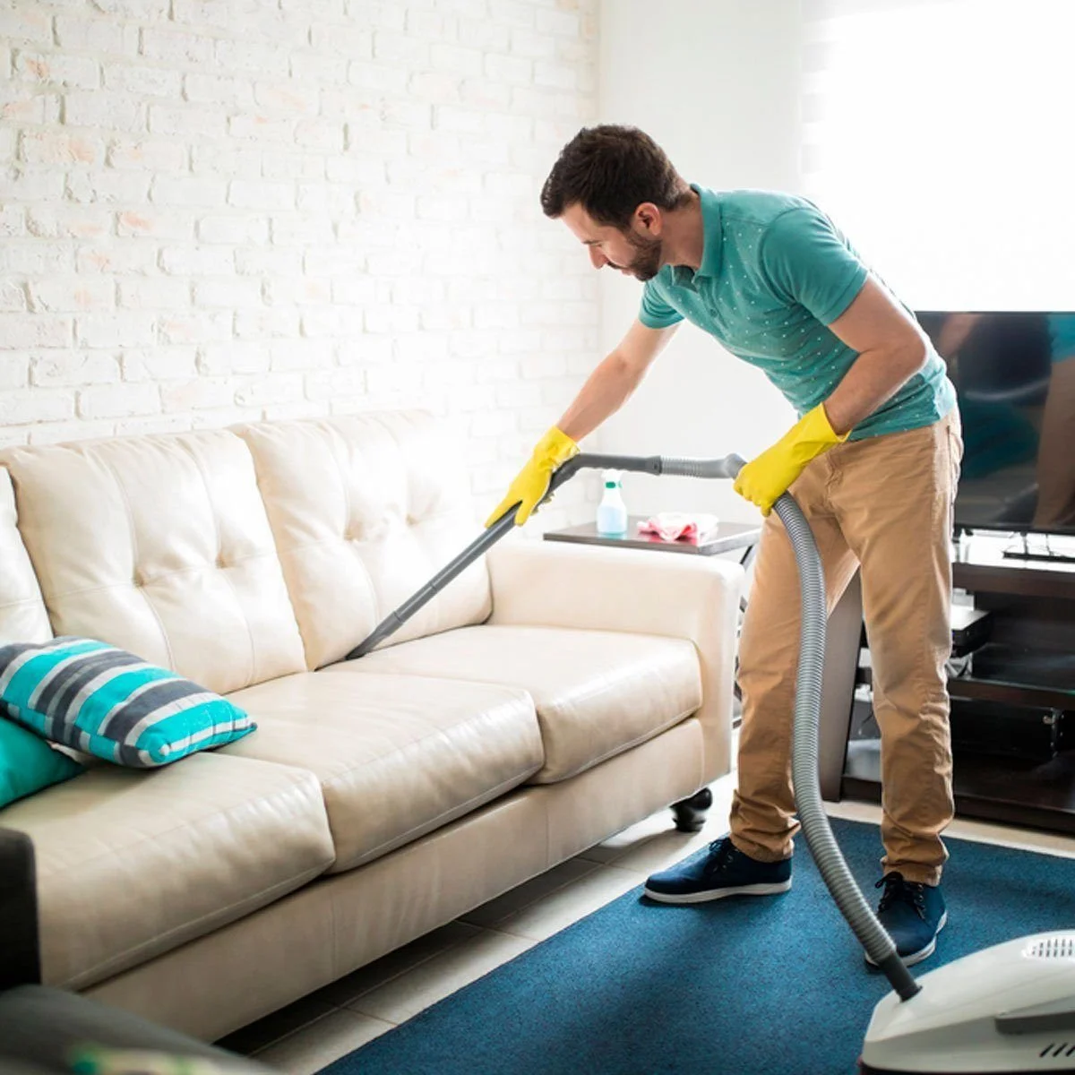 A man wearing yellow gloves vacuuming a cream-colored sofa in a living room with white brick walls and a blue carpet.