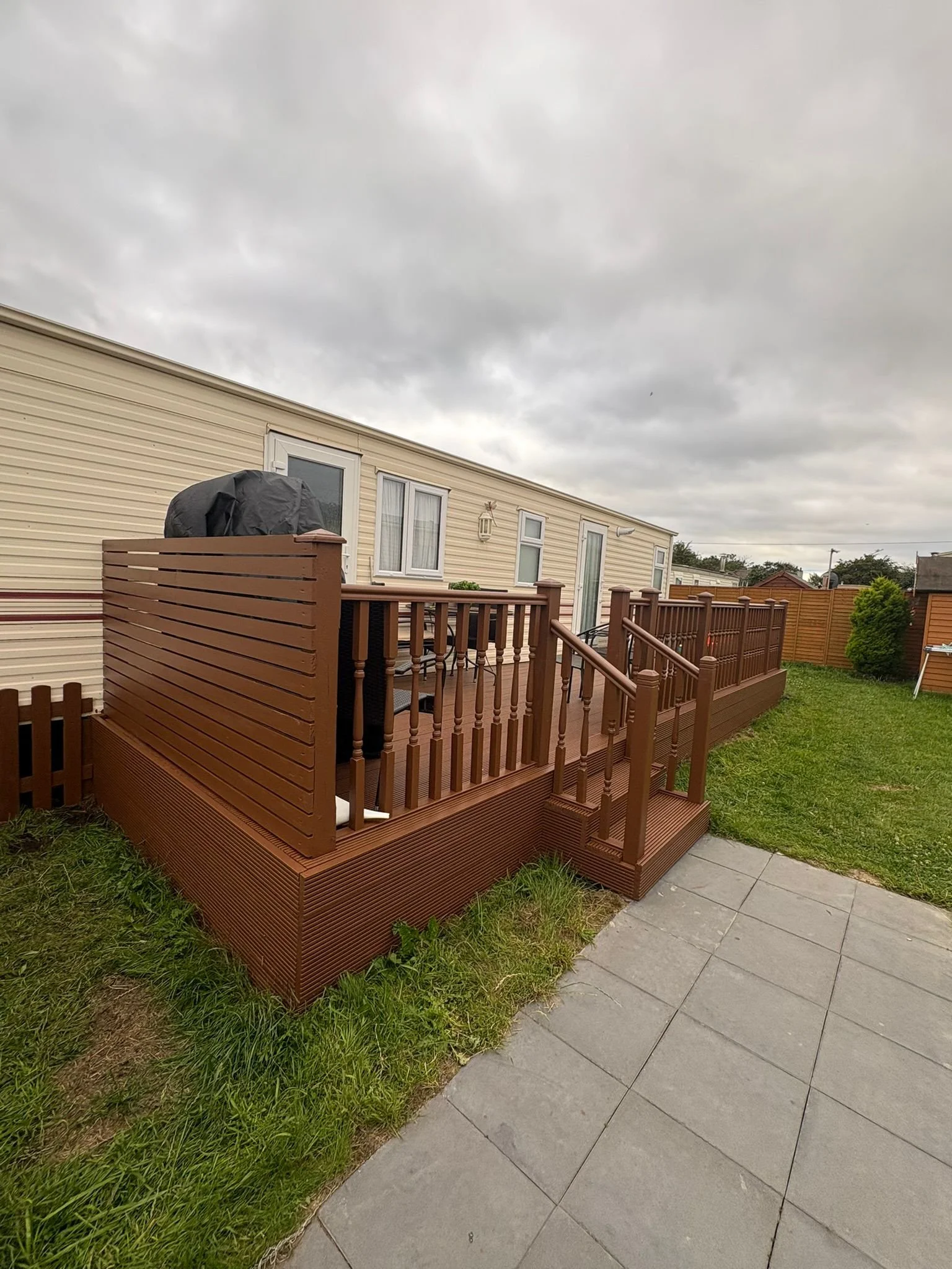 Backyard patio with newly installed brown wooden deck and railing, with an outdoor grill covered by a cover, adjacent to a cream-colored mobile home, and a small patch of grass and a paved area in the foreground, under overcast sky.