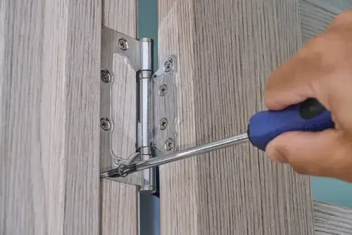 Hand using a screwdriver to tighten a door hinge on a wooden door.