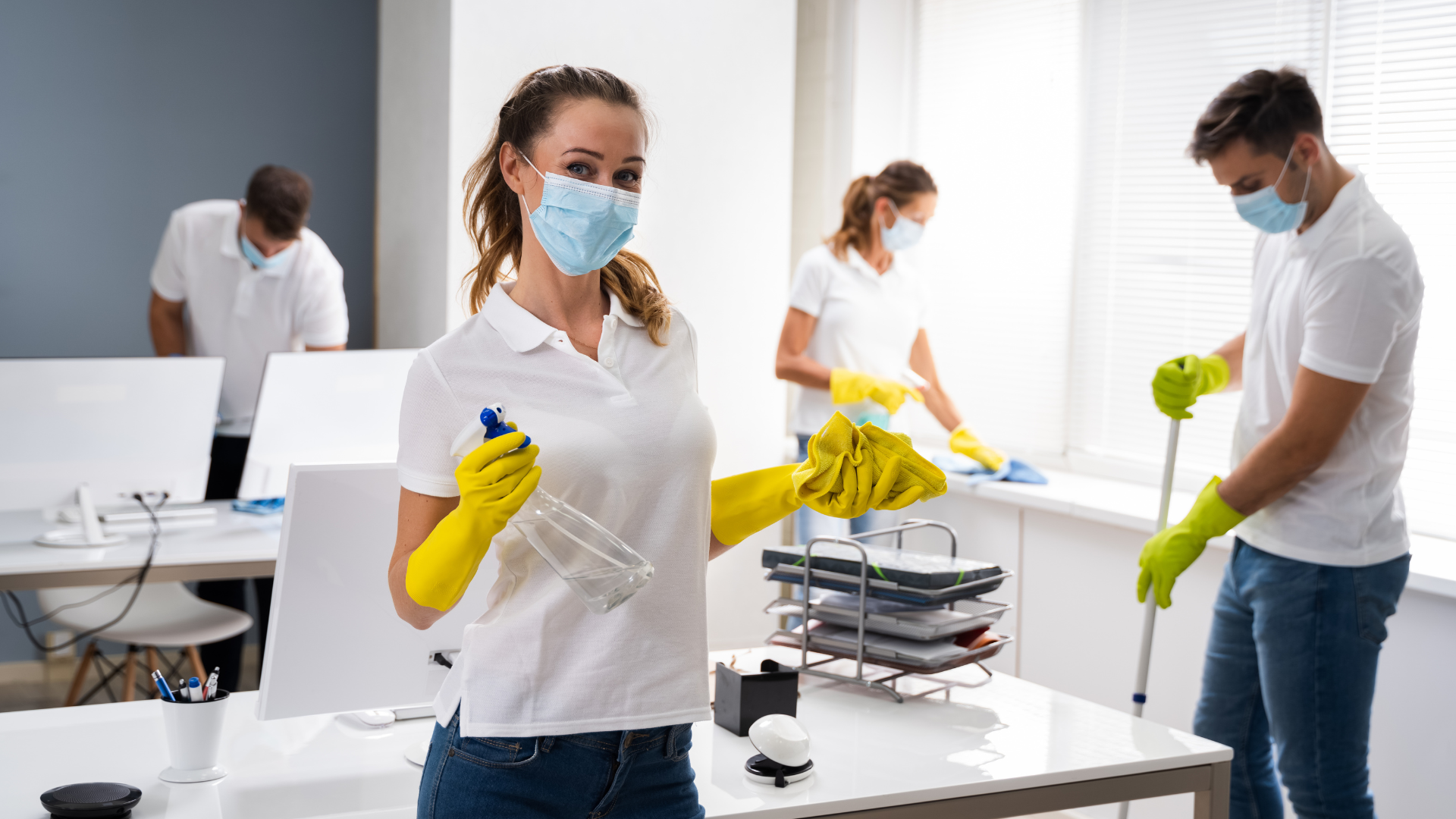 Group of four people in an office cleaning, wearing masks and yellow gloves. One woman in the foreground holds a spray bottle and a cleaning cloth, smiling at the camera, with two men and another woman in the background cleaning surfaces.