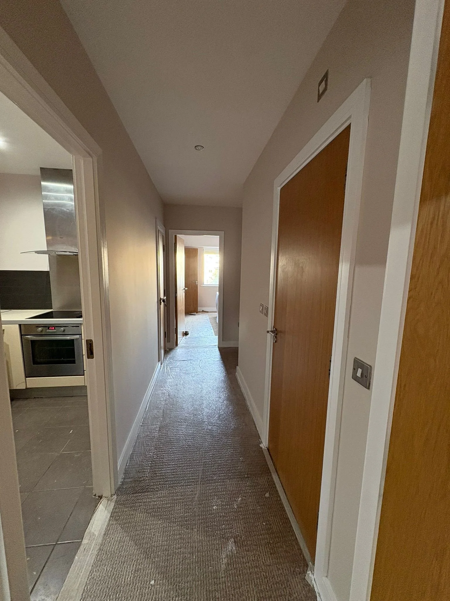 A hallway inside a house with beige walls, brown wooden doors, carpeted floor, and a view into a kitchen on the left and a room at the end of the hall with a window.