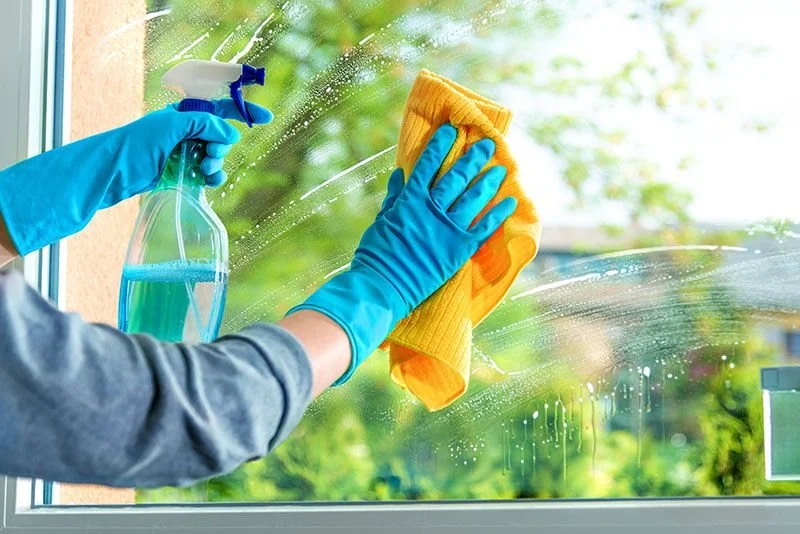 Person cleaning a window with a spray bottle and a yellow cloth, wearing blue gloves.