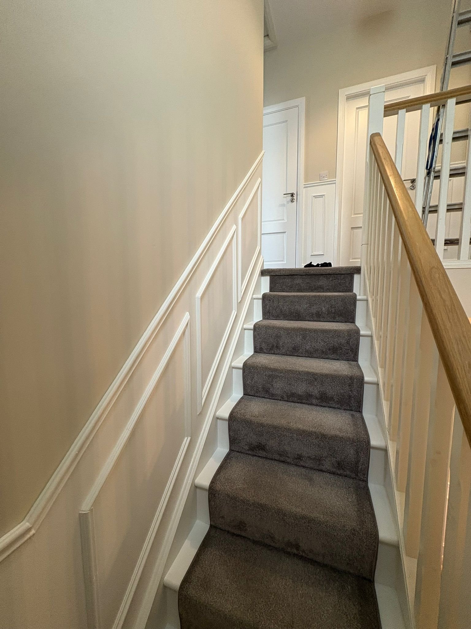 Inside a house staircase with gray carpeted stairs, beige walls, white wainscoting, and white doors at the top of the stairs.