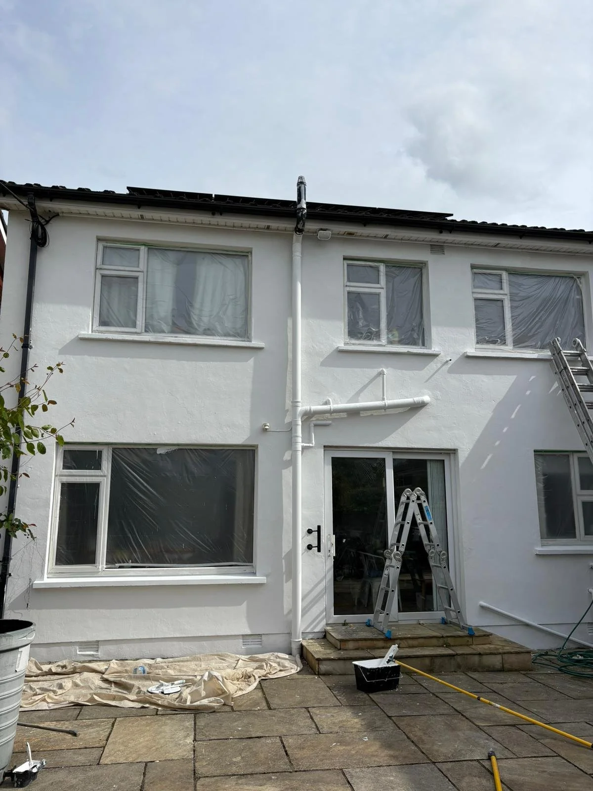 The back view of a white two-story house undergoing painting or cleaning, with two ladders leaning against the wall, window covers, and construction tools on the patio.