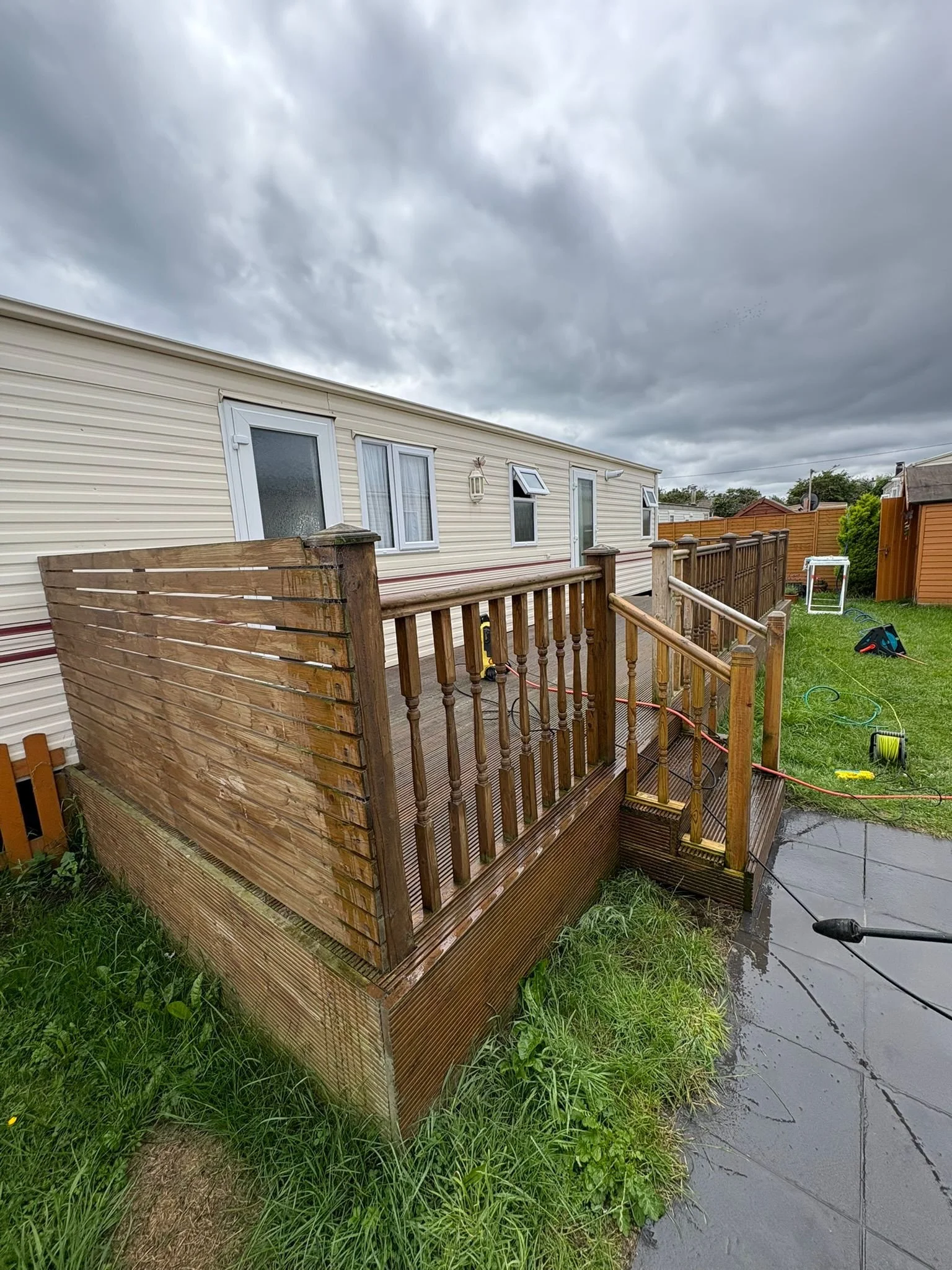 A small wooden deck with a railing attached to a mobile home, with a cloudy sky overhead. The deck is wet and has some gardening tools and equipment nearby on the grass and concrete patio.