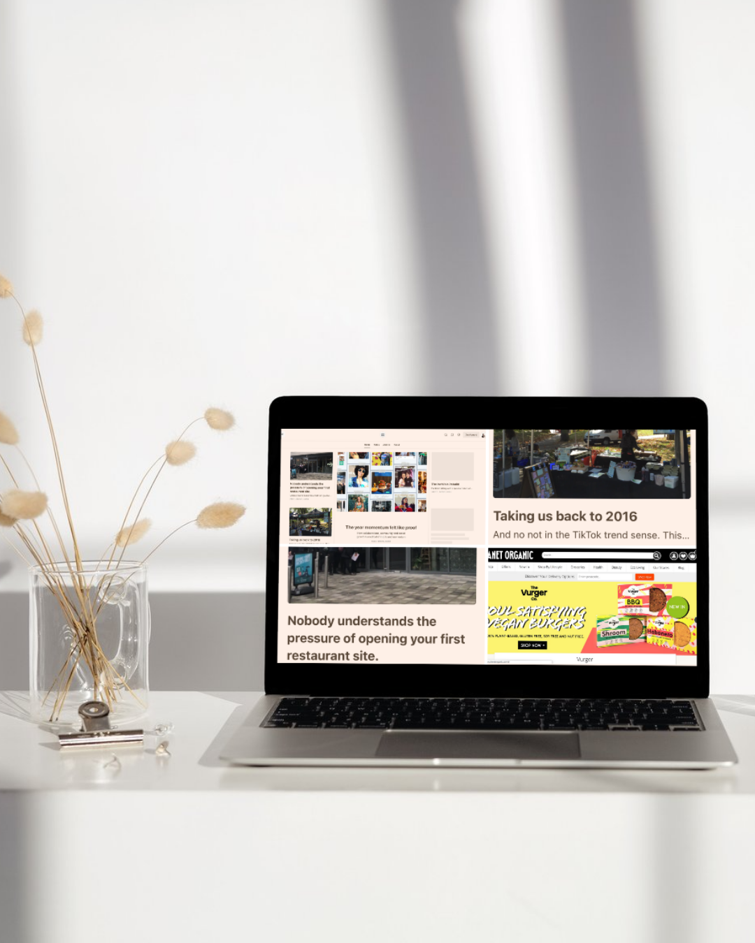 Open laptop on white desk displaying a website with articles and images, beside a clear glass vase with dried flowers, in a minimalist room with white walls and shadows.