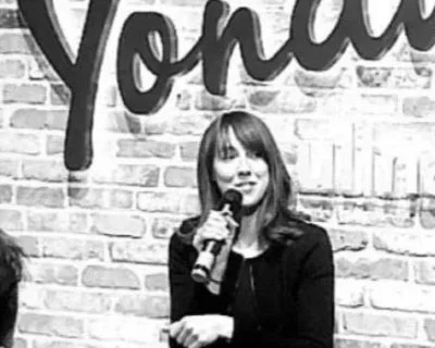 A woman with shoulder-length hair speaking into a microphone on stage, with a brick wall and partial word 'Yonder' in the background.