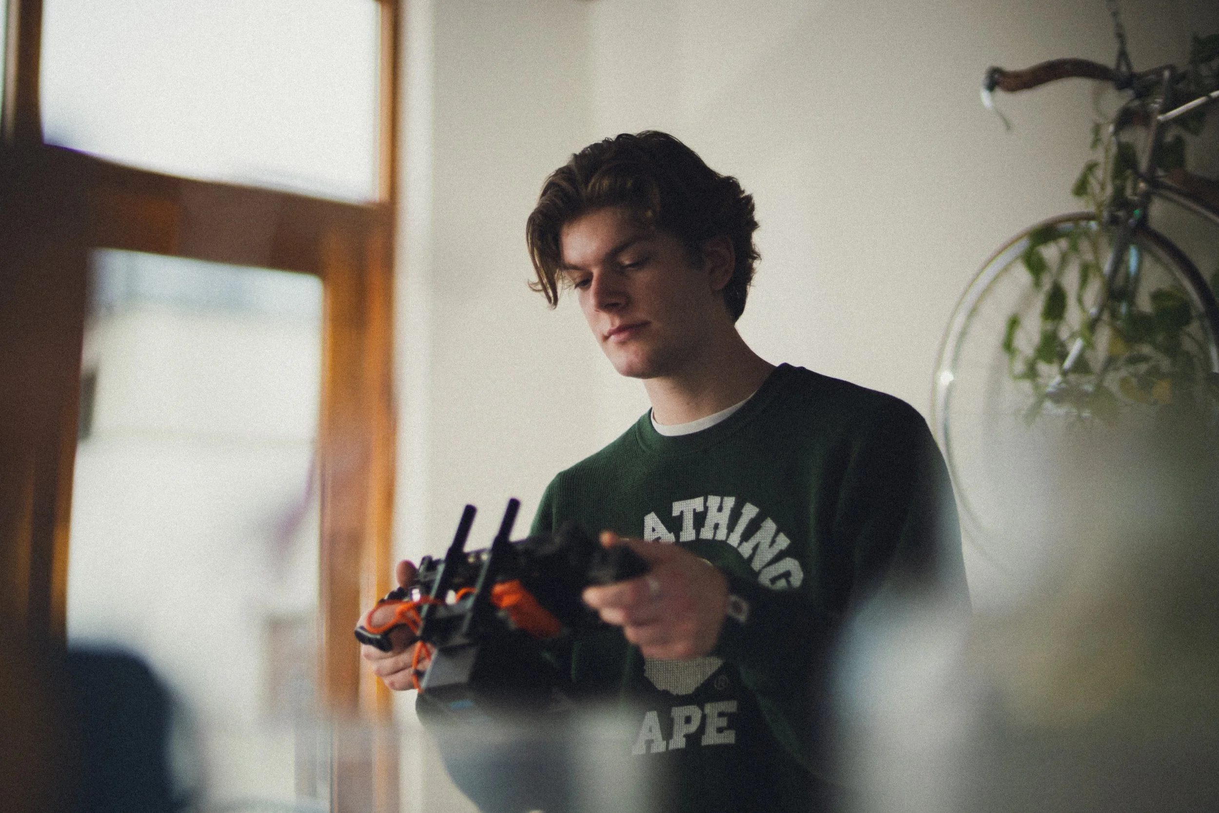 A young man with brown hair wearing a green sweatshirt, holding a remote control drone indoors, with a bookshelf and bicycle in the background.