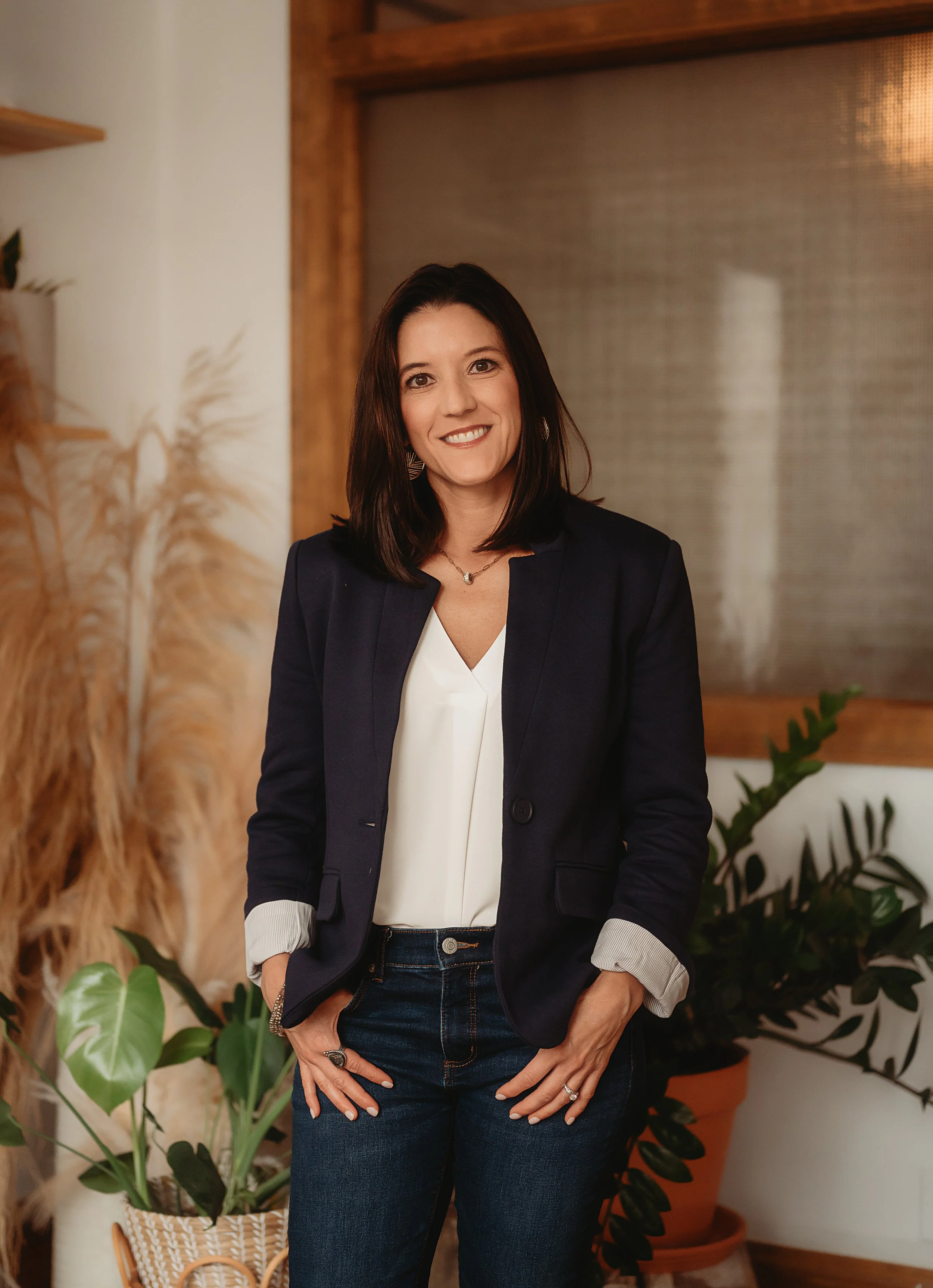 A woman with shoulder-length brown hair, wearing a black blazer and white shirt, standing in a room with plants and wooden accents, smiling at the camera.