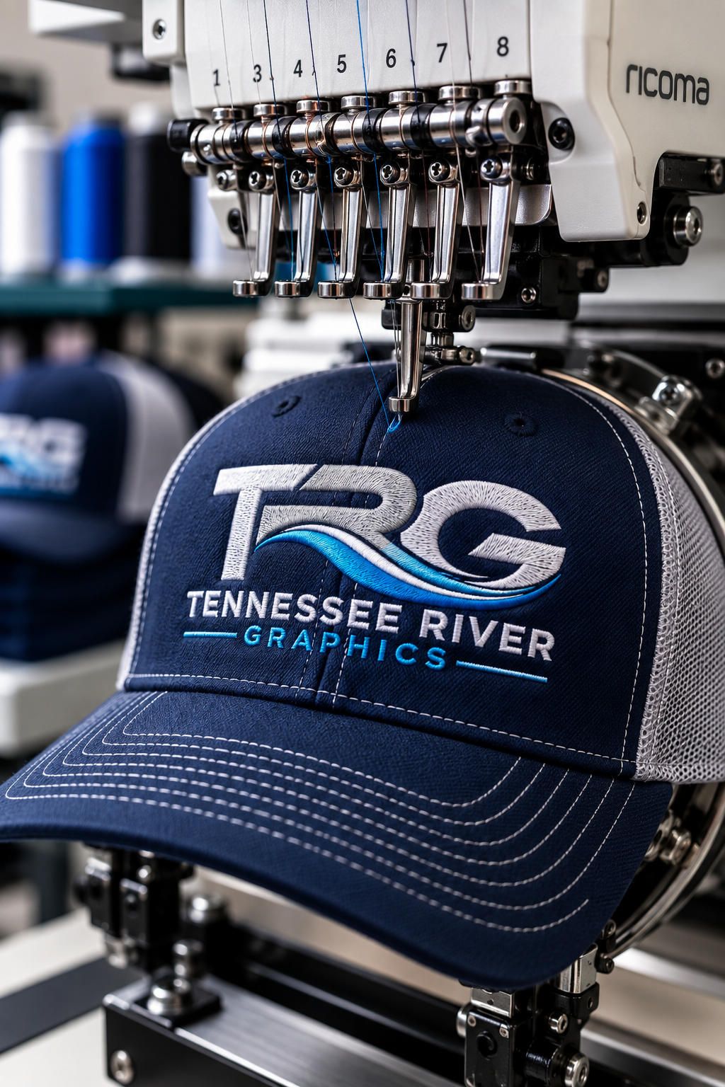 A embroidery machine stitching a logo onto a navy blue cap. The logo features the text 'Tennessee River Graphics' with a stylized wave design, and the embroidery process is in progress.