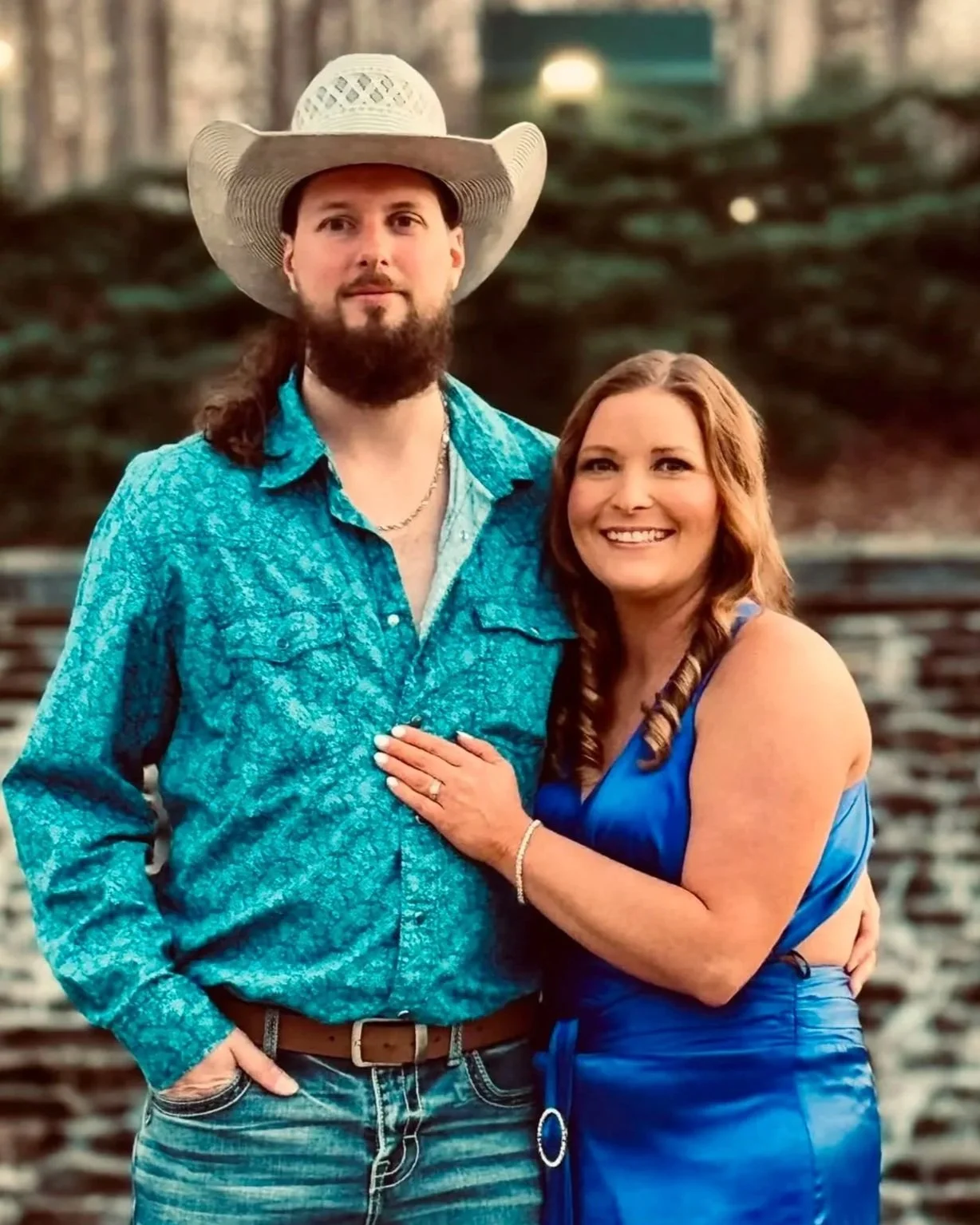 A man and woman posing outdoors, the man with a beard and wearing a cowboy hat, a blue patterned shirt, and jeans, and the woman with shoulder-length hair, wearing a blue dress, smiling.