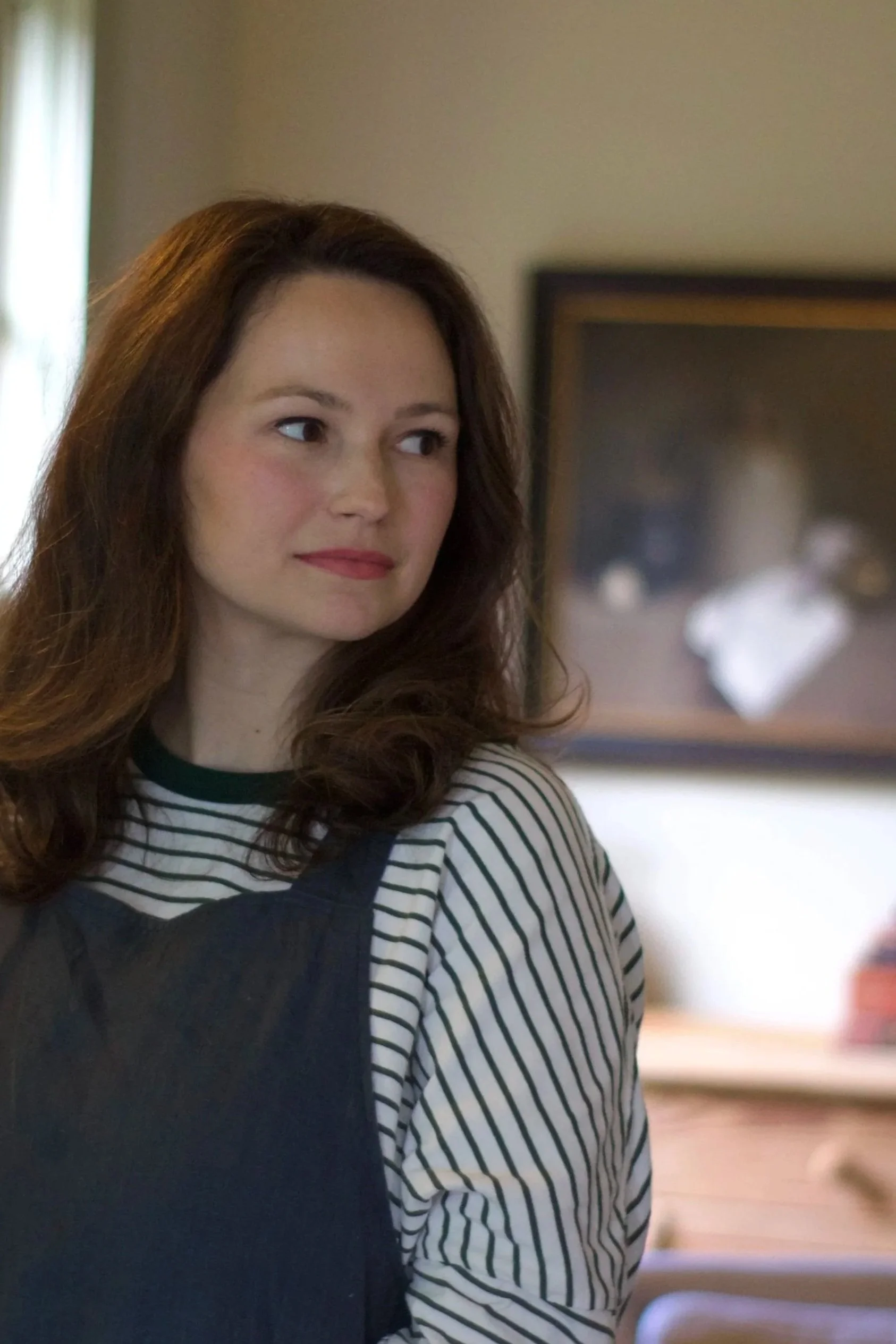 A woman with long, wavy brown hair and wearing a striped shirt and black apron, standing indoors near a window with green curtains, and a blurred framed picture in the background.