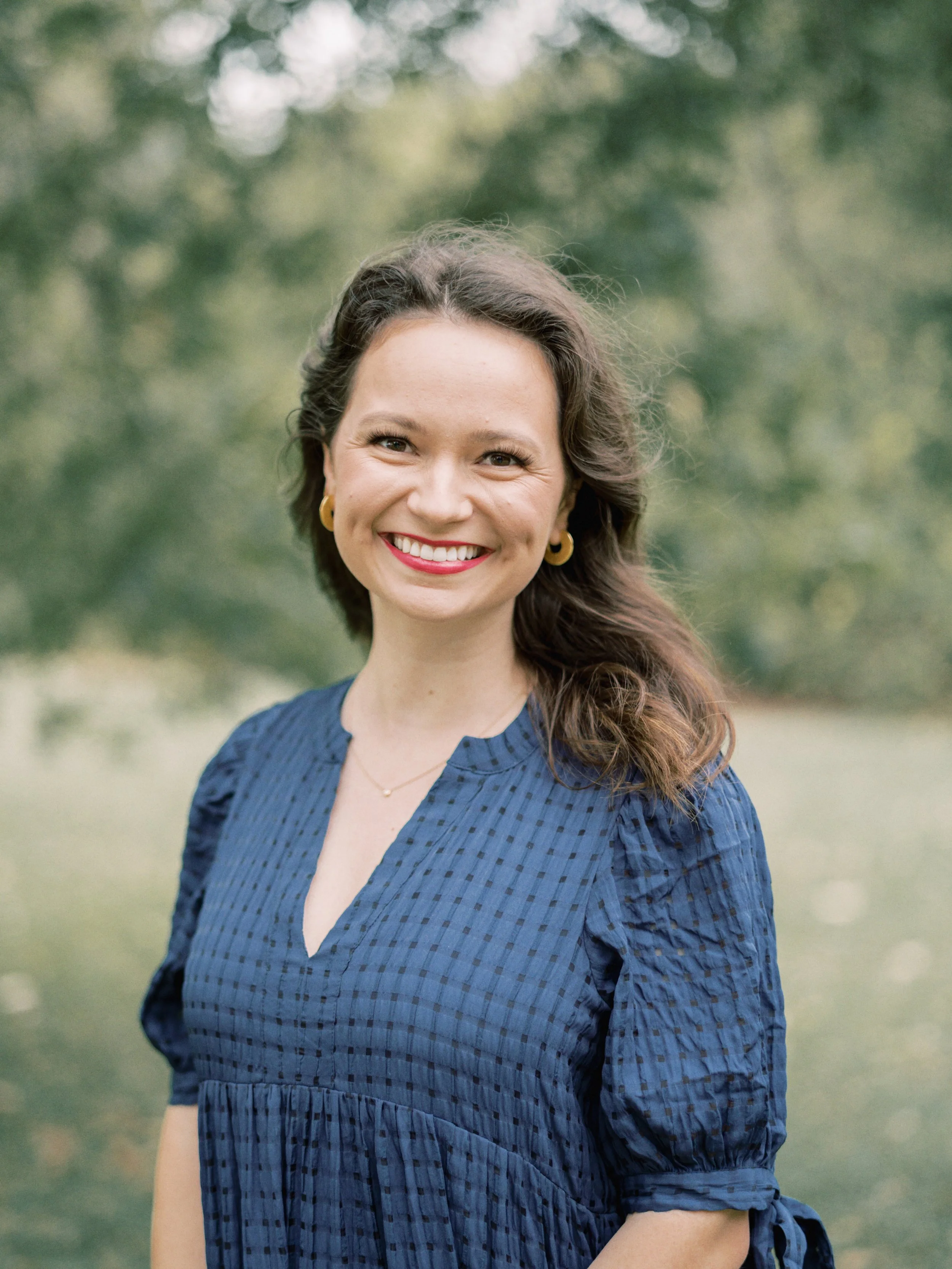 A woman smiling outdoors with trees in the background, wearing a blue dress and gold jewelry.