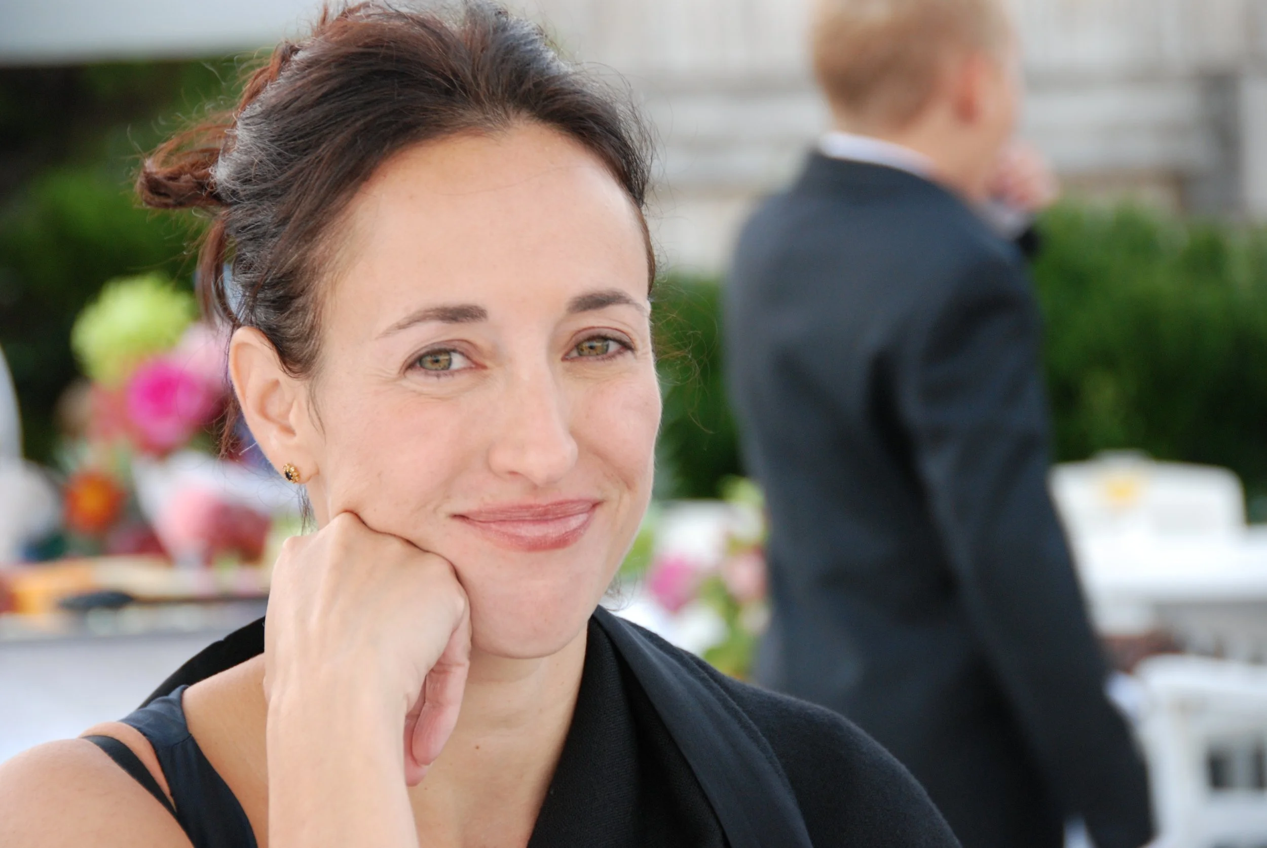 Julie Farris landscape designer with  with brown hair, gold earrings, and light makeup resting her chin on her hand at an outdoor event, with a blurred man in a suit and colorful flowers in the background.