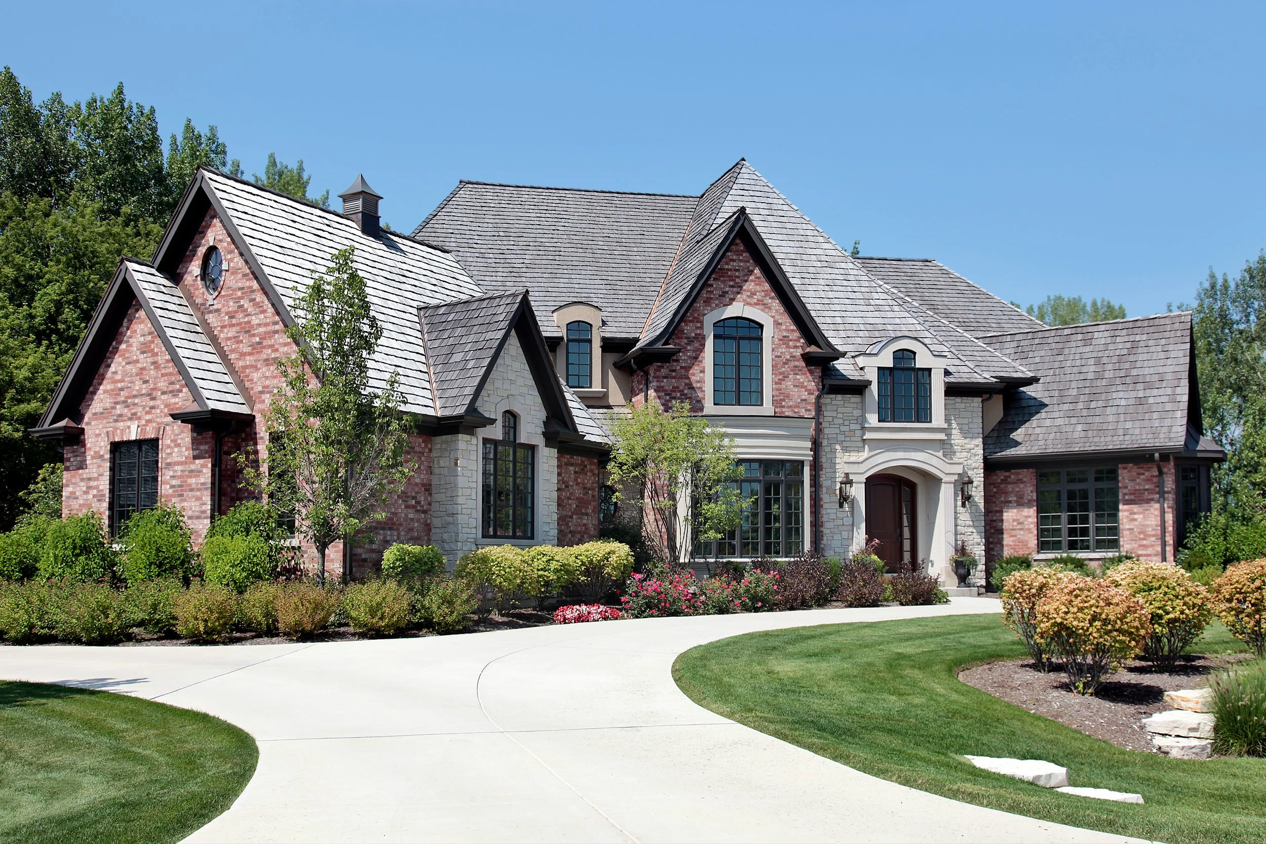 A large, elegant house with brick and stone exterior, multiple gabled roofs, and landscaped front yard with trees, bushes, and a curved driveway.