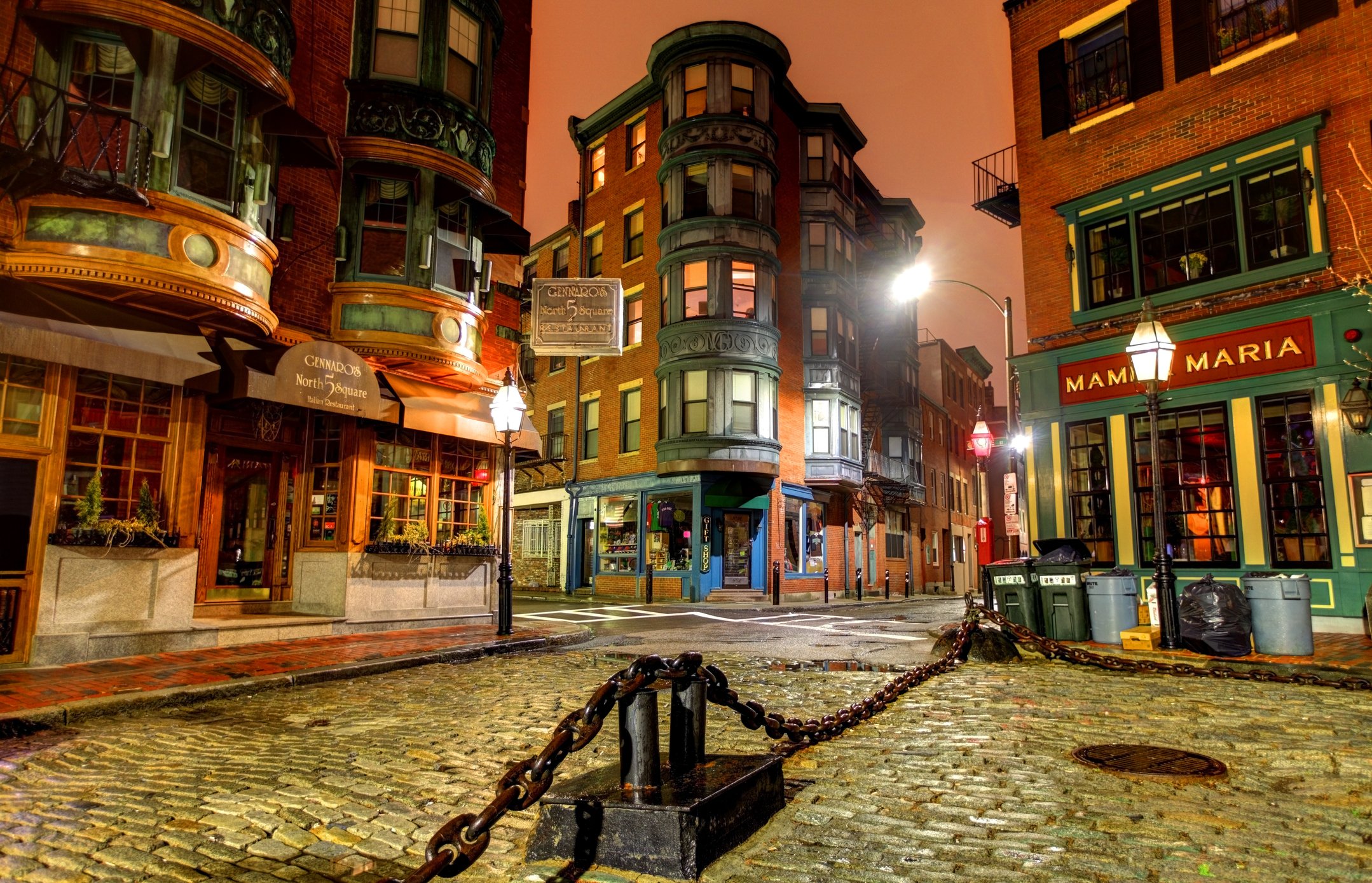 Street view of Boston's North End at night with cobblestone pavement, historic brick buildings, chain barrier, and illuminated signs for restaurants including "Gennaro's North Square" and "Mamma Maria."