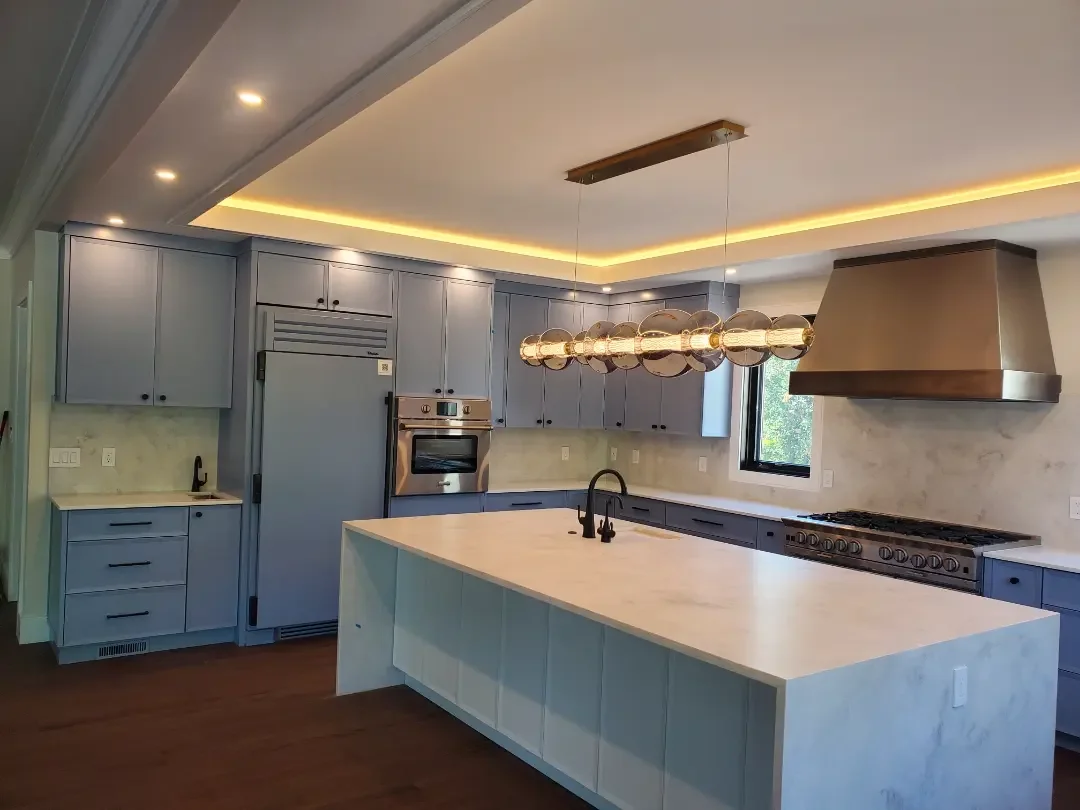 Modern kitchen with light blue cabinets, a large white island with a black faucet, stainless steel double oven, and gas cooktop. There is a large overhead light fixture with multiple glass globes, a window near the cooktop, and recessed lighting alon